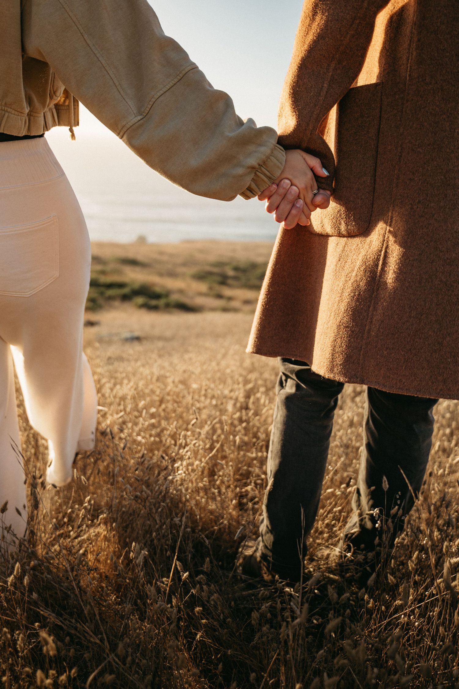 A couple holds hands while walking through tall dry grass toward the ocean, wearing warm autumn-toned coats at sunset.