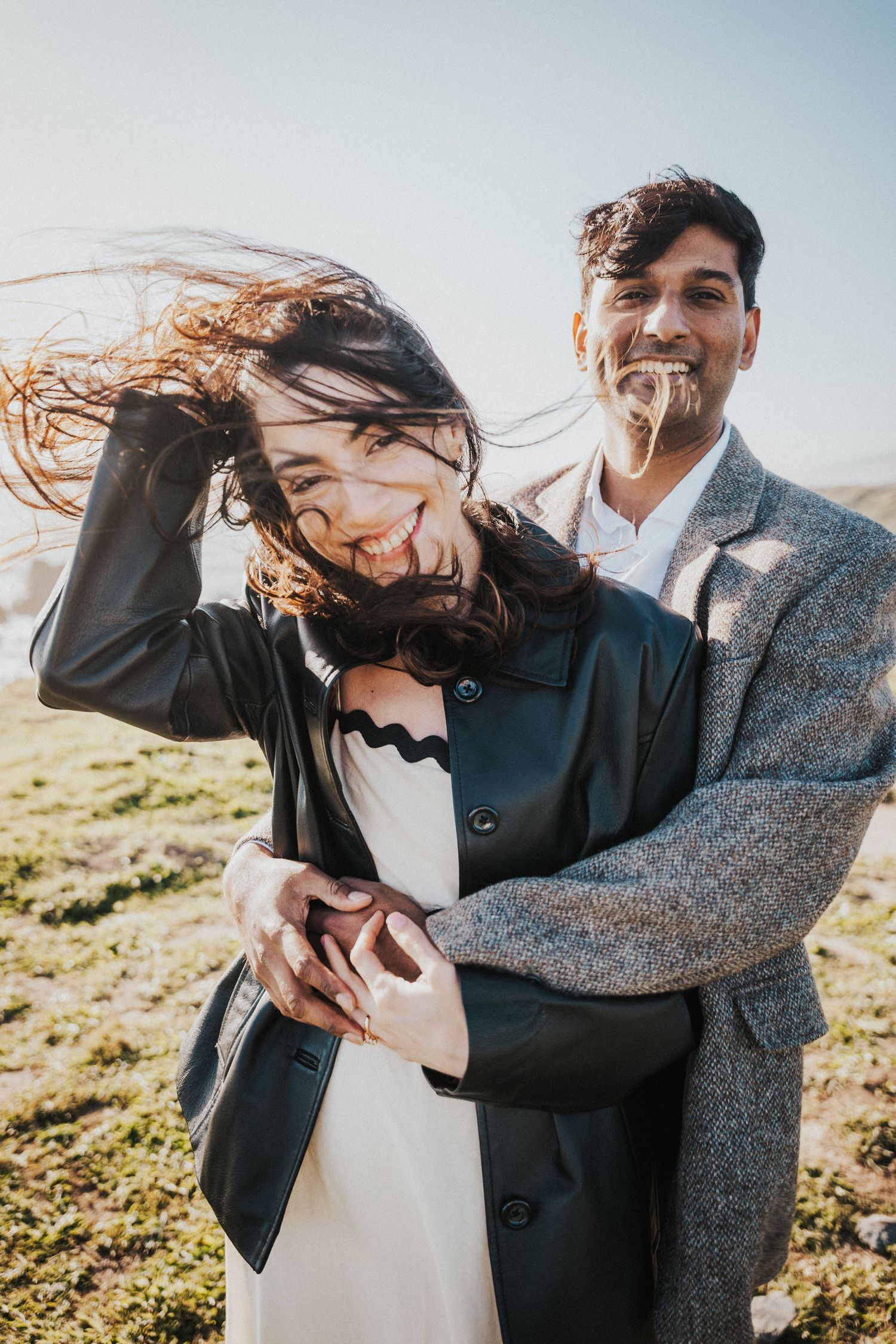 Couple embracing outdoors on a windy day, woman laughing in black leather jacket, man in grey blazer holding her.