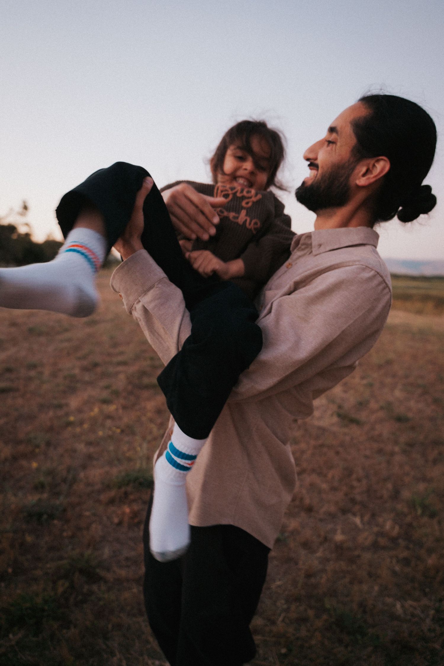 A bearded man joyfully lifts a laughing child in an open field during golden hour.