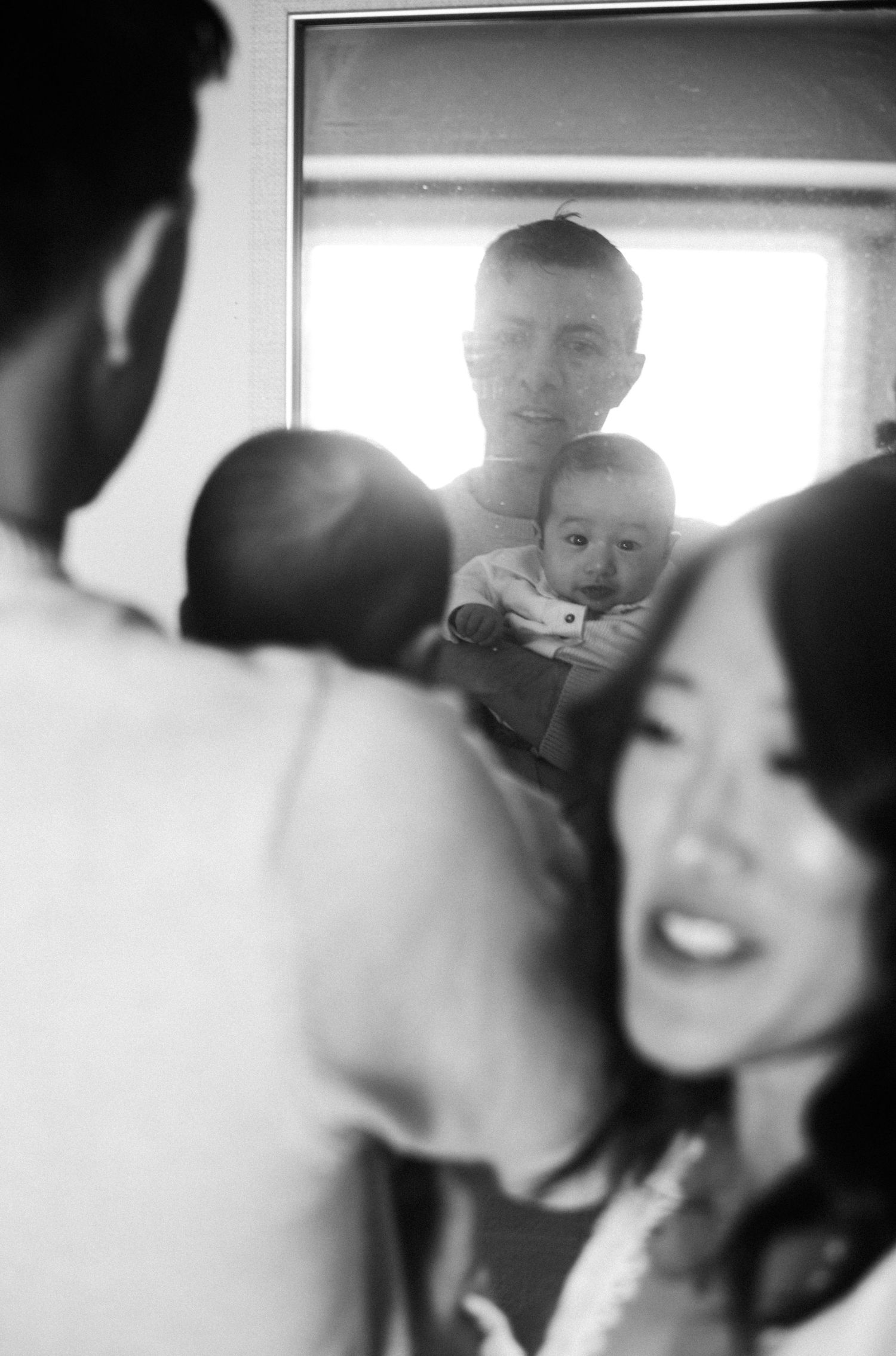 Black and white mirror reflection of a father holding a newborn baby with a smiling mother in the foreground.