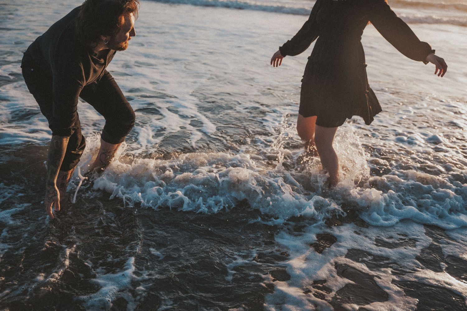 Two people in dark clothes splashing through ocean waves at the beach during golden hour.Photography by The Y.K. Studio.