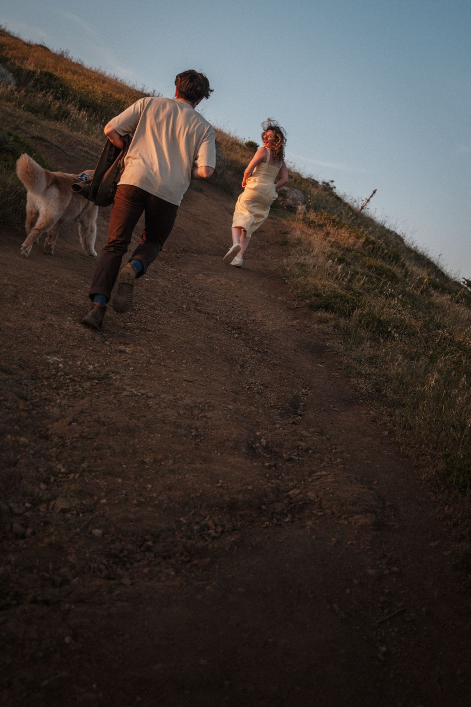 Couple in wedding attire running up a dirt path at sunset with a dog beside them. Photography by The Y.K. Studio.