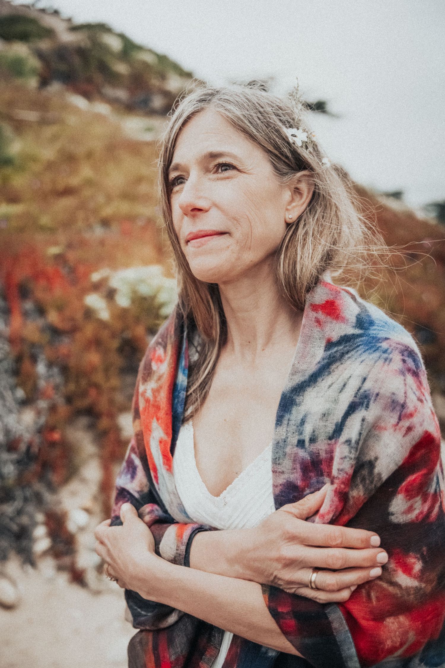 Middle-aged woman with flower in hair wrapped in colorful tie-dye shawl gazing thoughtfully at coastal landscape.