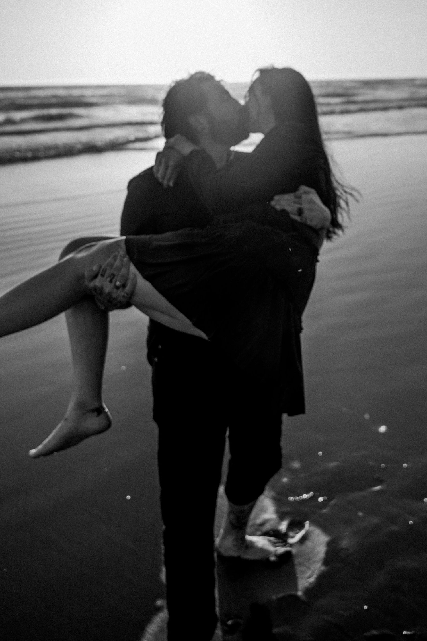 Man carrying woman while kissing on beach at sunset in black and white.