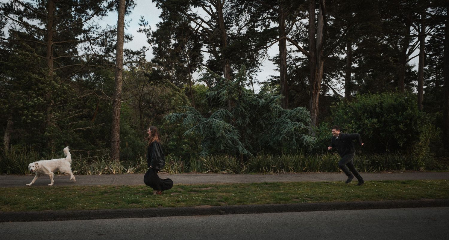 Two people in black clothing run across a grassy path with a white dog near tall trees.