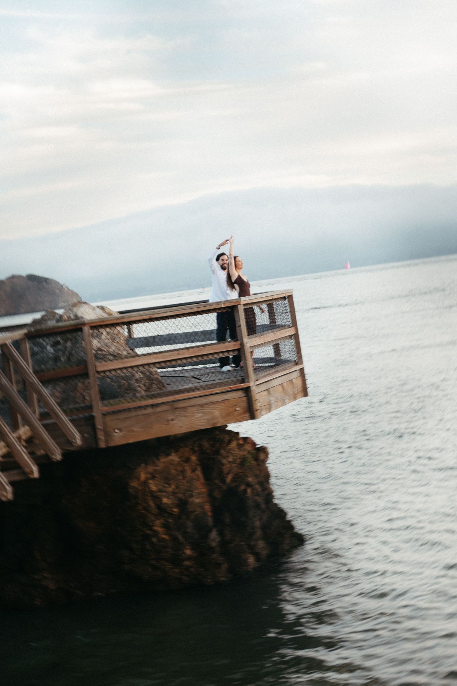 A couple poses on a wooden cliffside overlook above the ocean on a cloudy day.