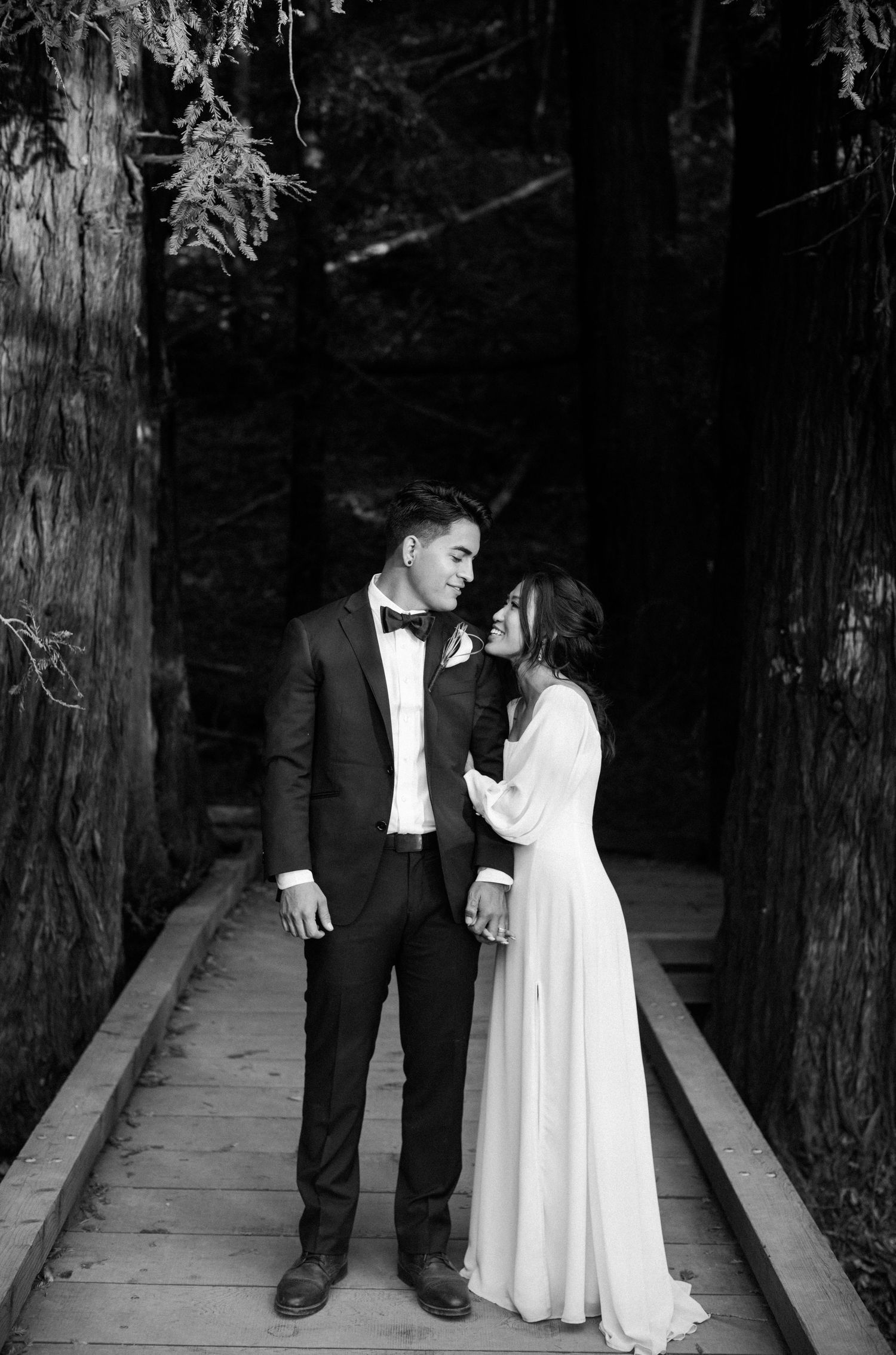 Black and white photo of a couple in wedding attire sharing an intimate moment on a wooden path surrounded by tall trees.