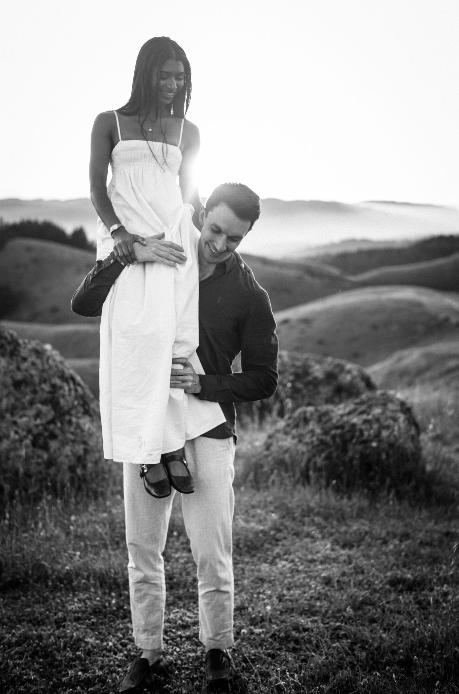 Black and white photo of man lifting woman on his shoulders in scenic rolling hills landscape at dusk.