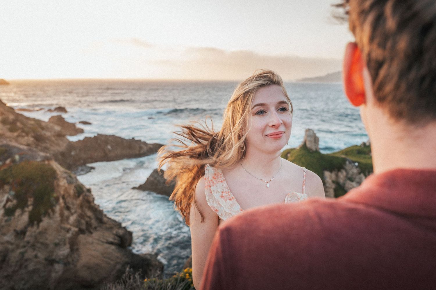 Smiling woman with windswept hair gazes at partner during romantic coastal sunset session.