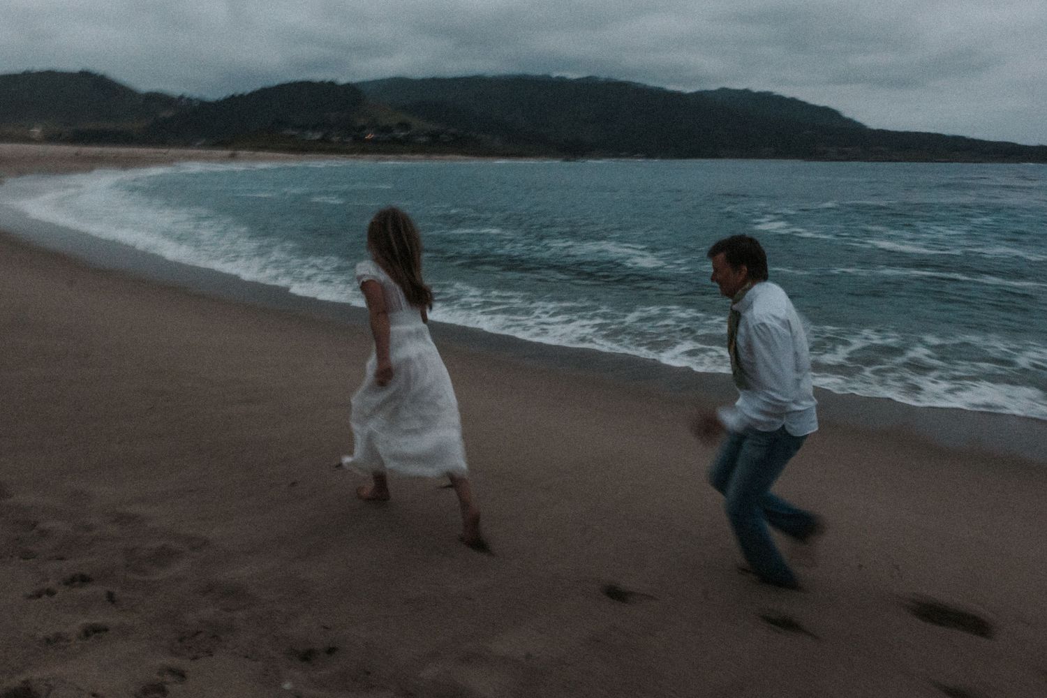 Couple running playfully on a sandy beach with ocean waves and misty mountains in the background.Photography by The Y.K. Studio.