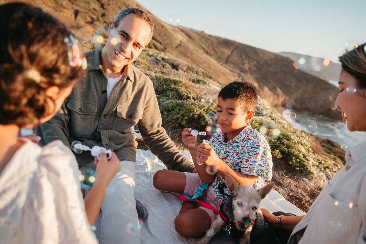 Family with dog enjoying a picnic on a scenic hillside, sharing marshmallows and smiling together outdoors.