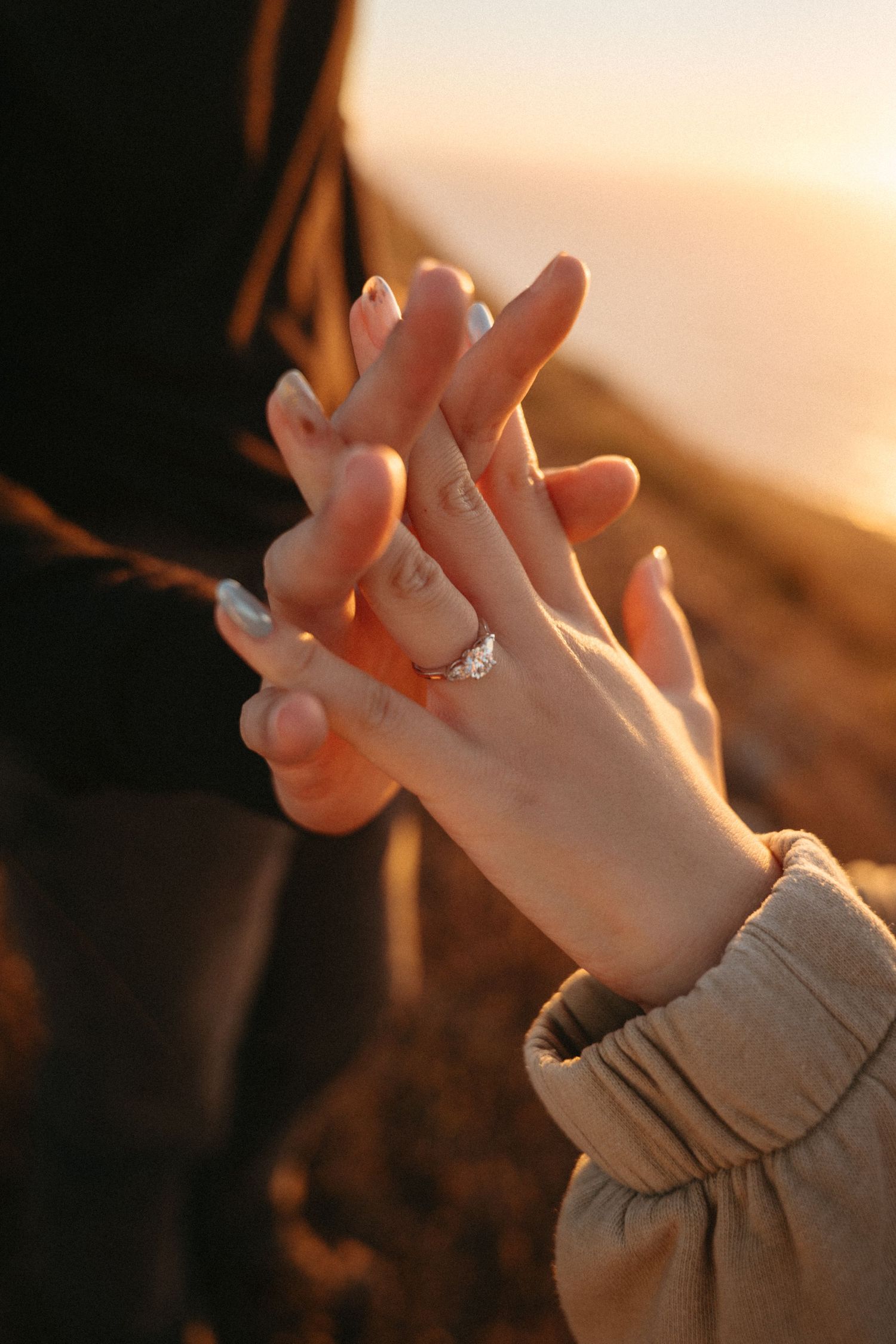 Two hands with interlaced fingers and a silver engagement ring glow in golden sunset light against a blurred coastal hillside.