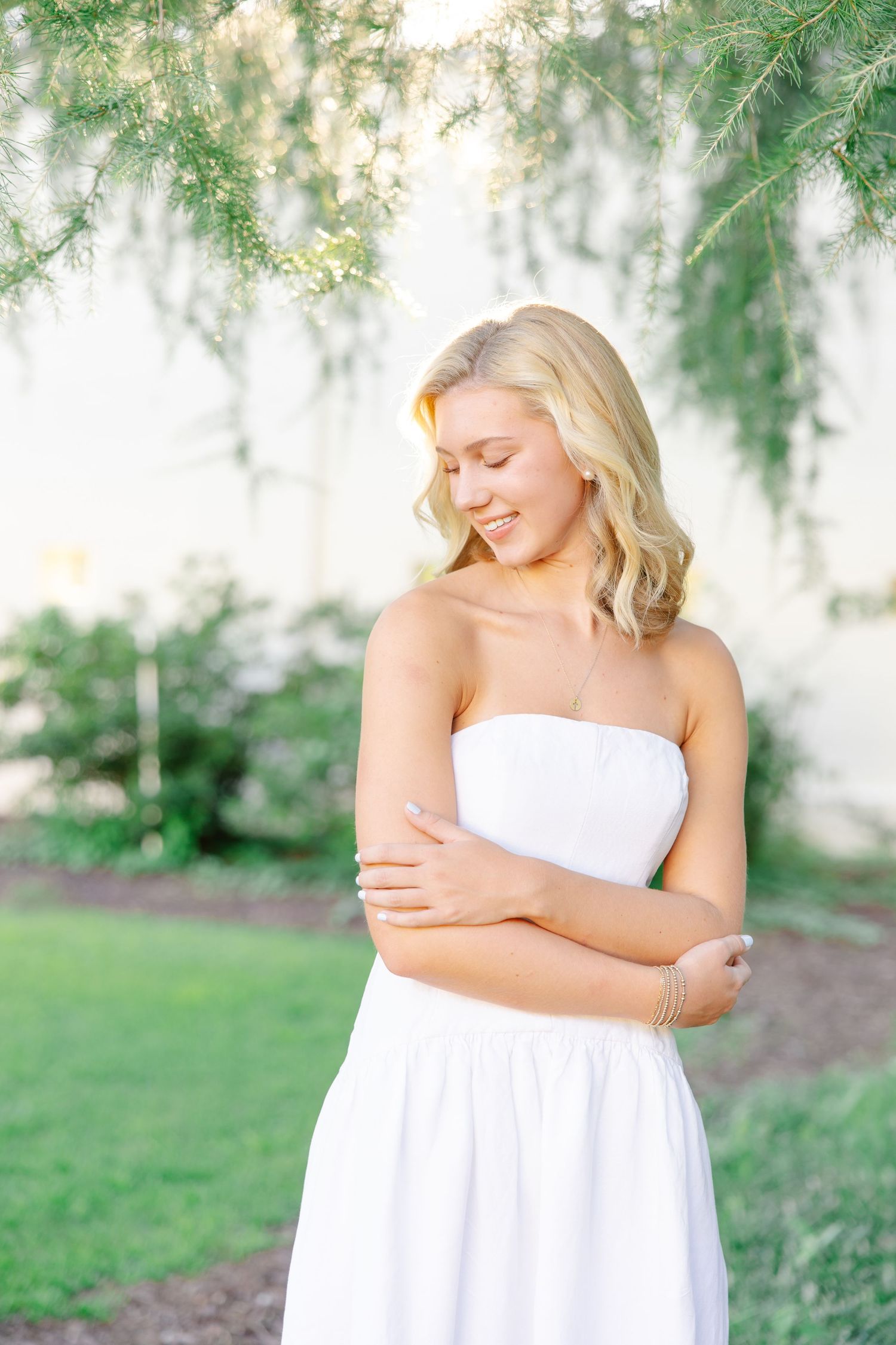 Blonde woman in white strapless dress hugging herself under green tree branches outdoors.