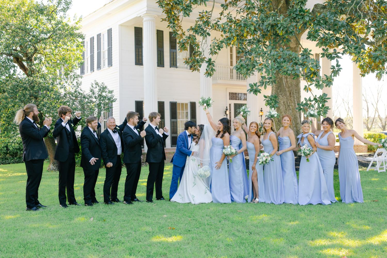Wedding party celebrating outdoors with bridesmaids in periwinkle gowns and groomsmen in black tuxedos before a white plantation home.