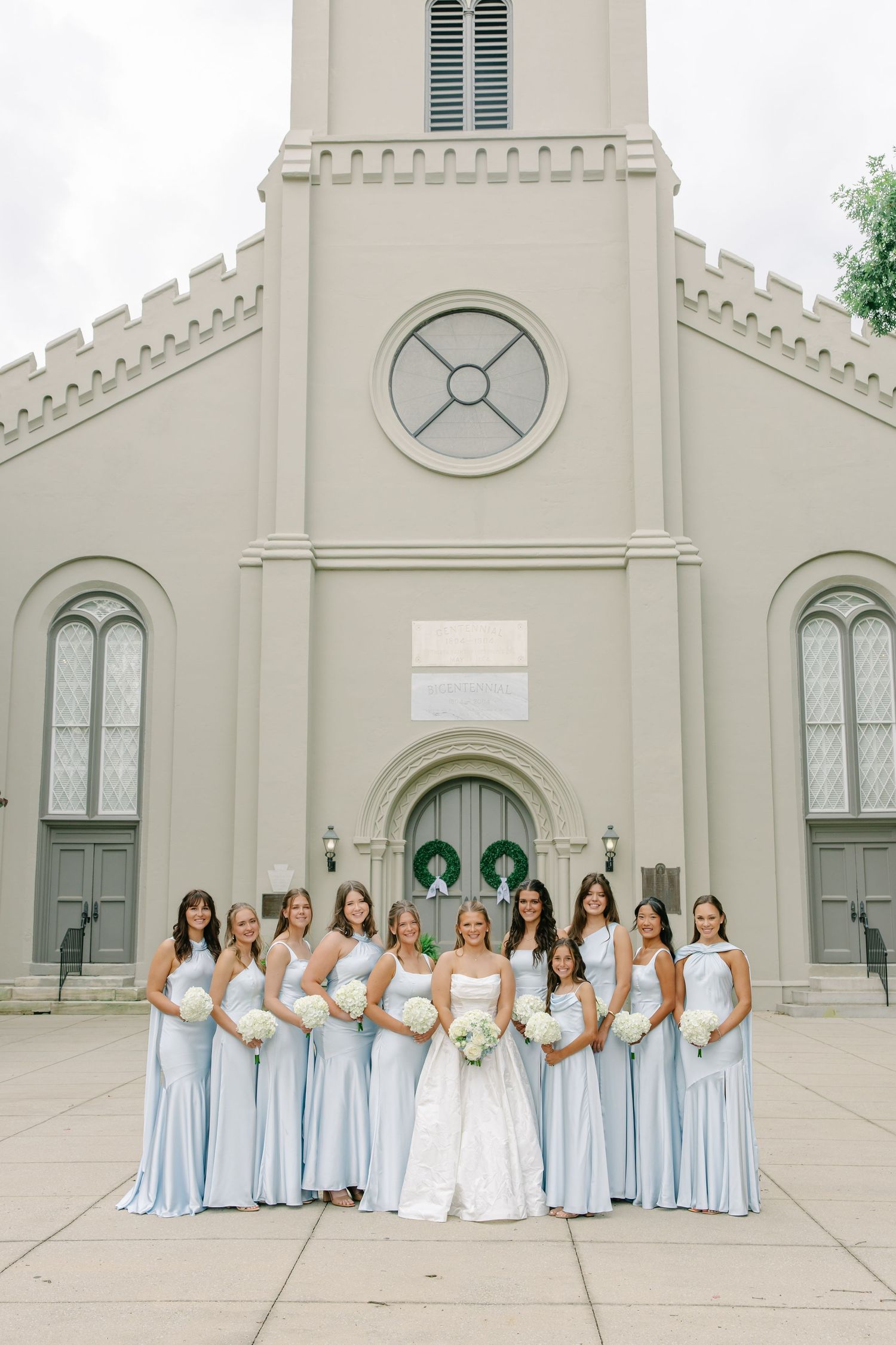 Bride in white ball gown stands with bridesmaids in pale blue dresses outside a classic stone church.