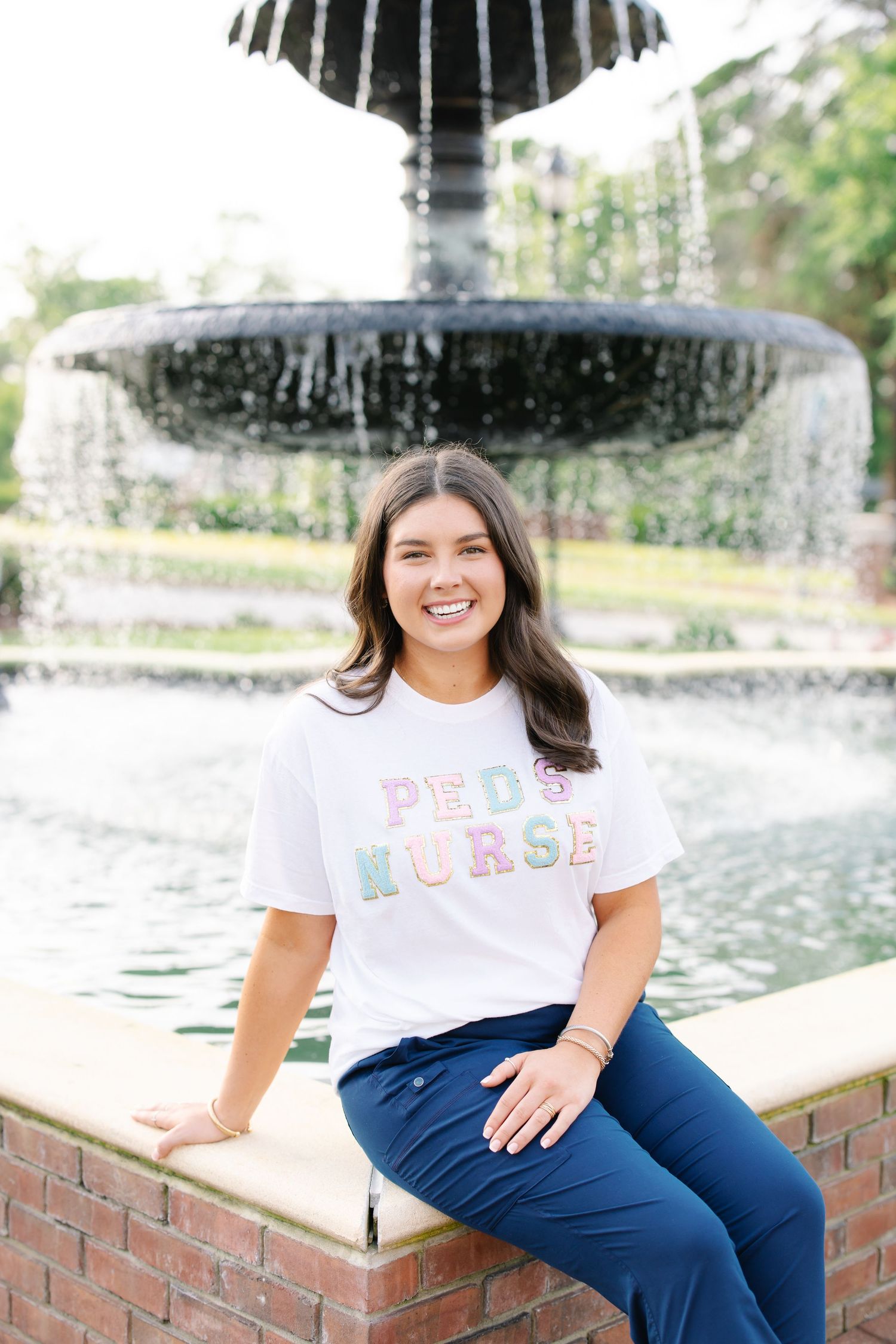 Smiling future nurse in white PhD Nurse t-shirt sitting by a decorative campus fountain.