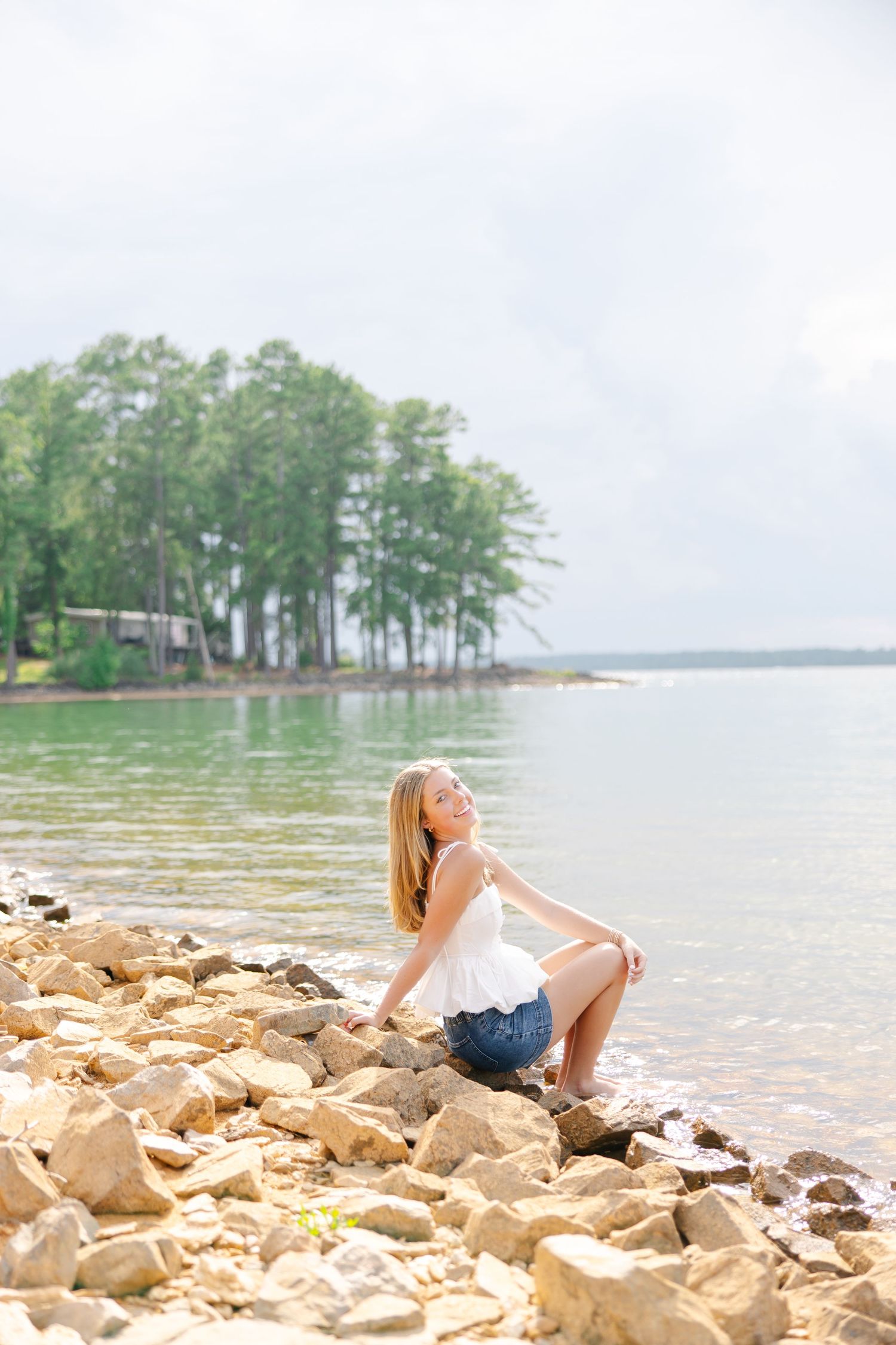 Blonde teen in white top and denim shorts sitting on rocky shoreline with green lake and trees behind.