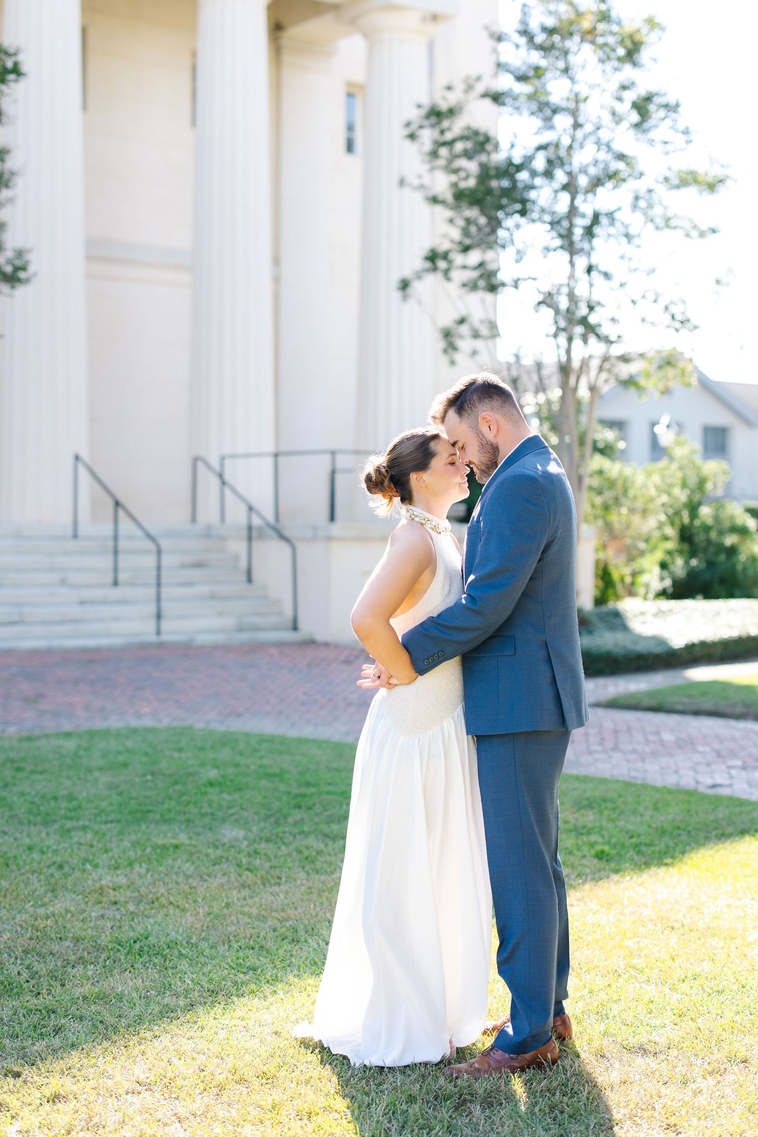 Bride and groom embracing on a sunny lawn in front of a white columned building after their ceremony.