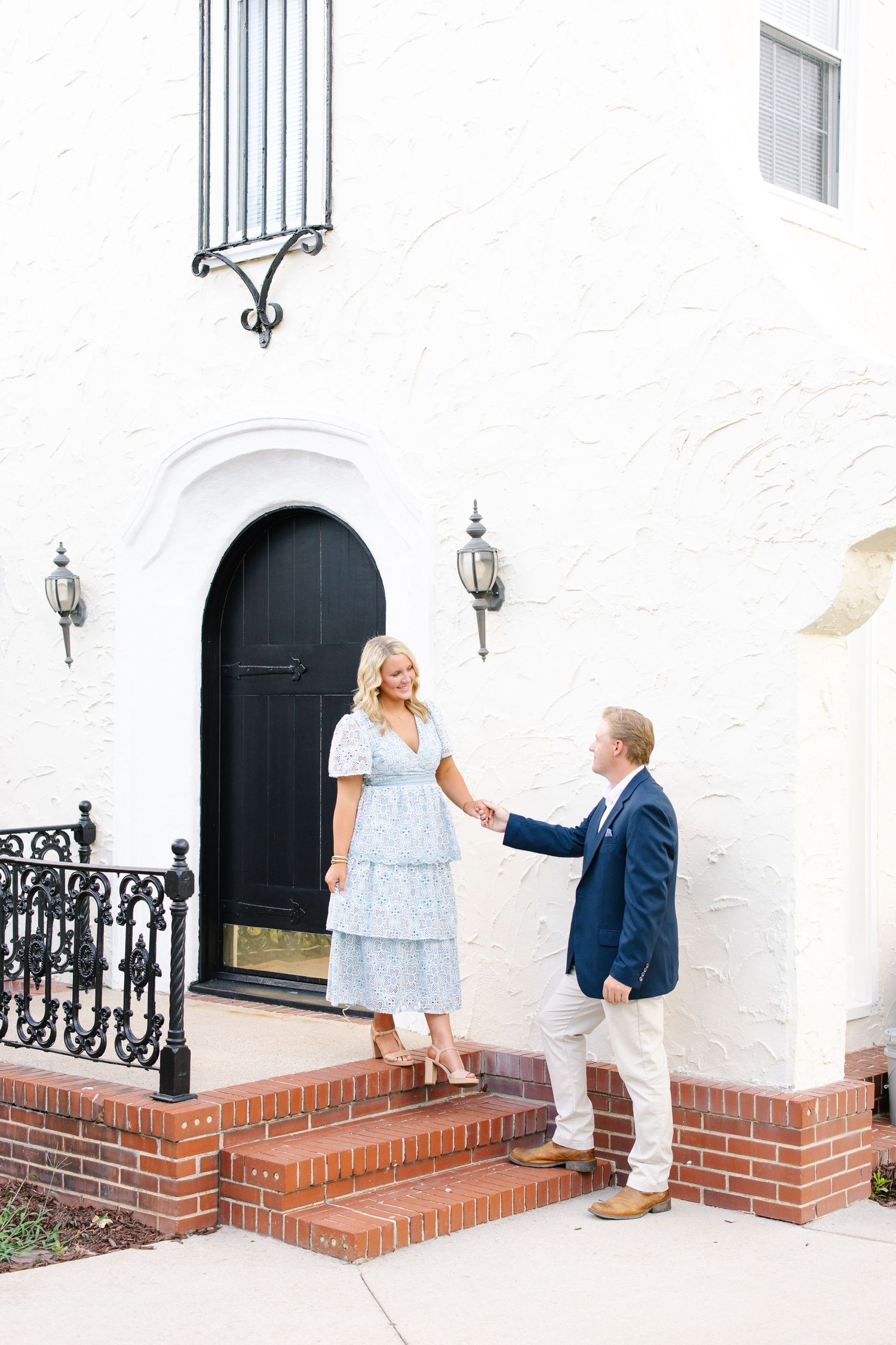 Couple holding hands on brick steps outside white stucco building with black arched door.