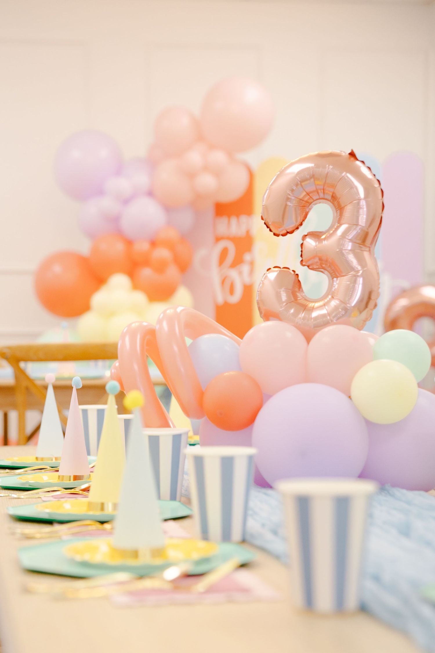 Rose gold number 3 balloon centerpiece with pastel balloon arch and striped party hats on birthday table.