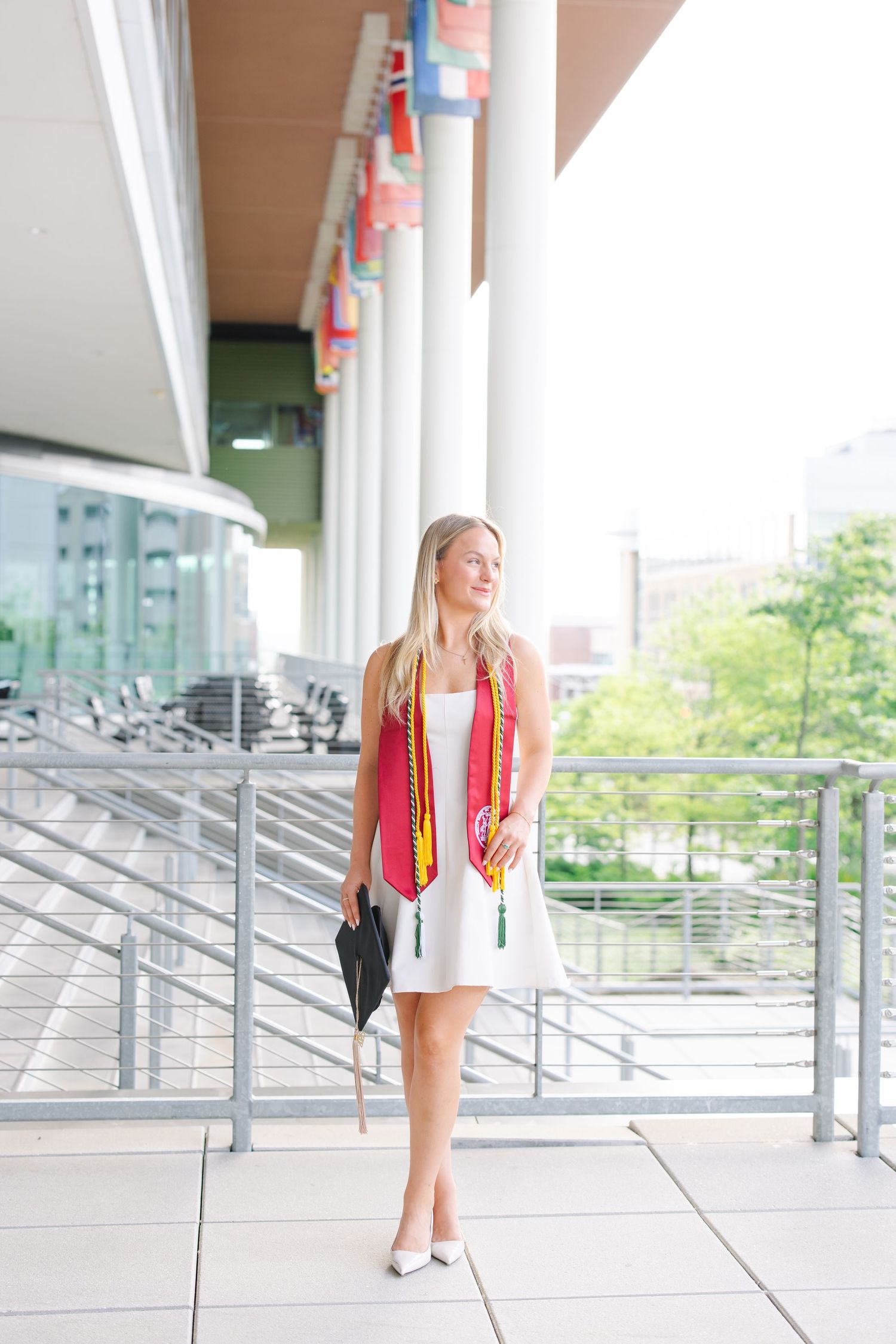 Blonde college graduate in white dress holding cap with honor stoles at university.