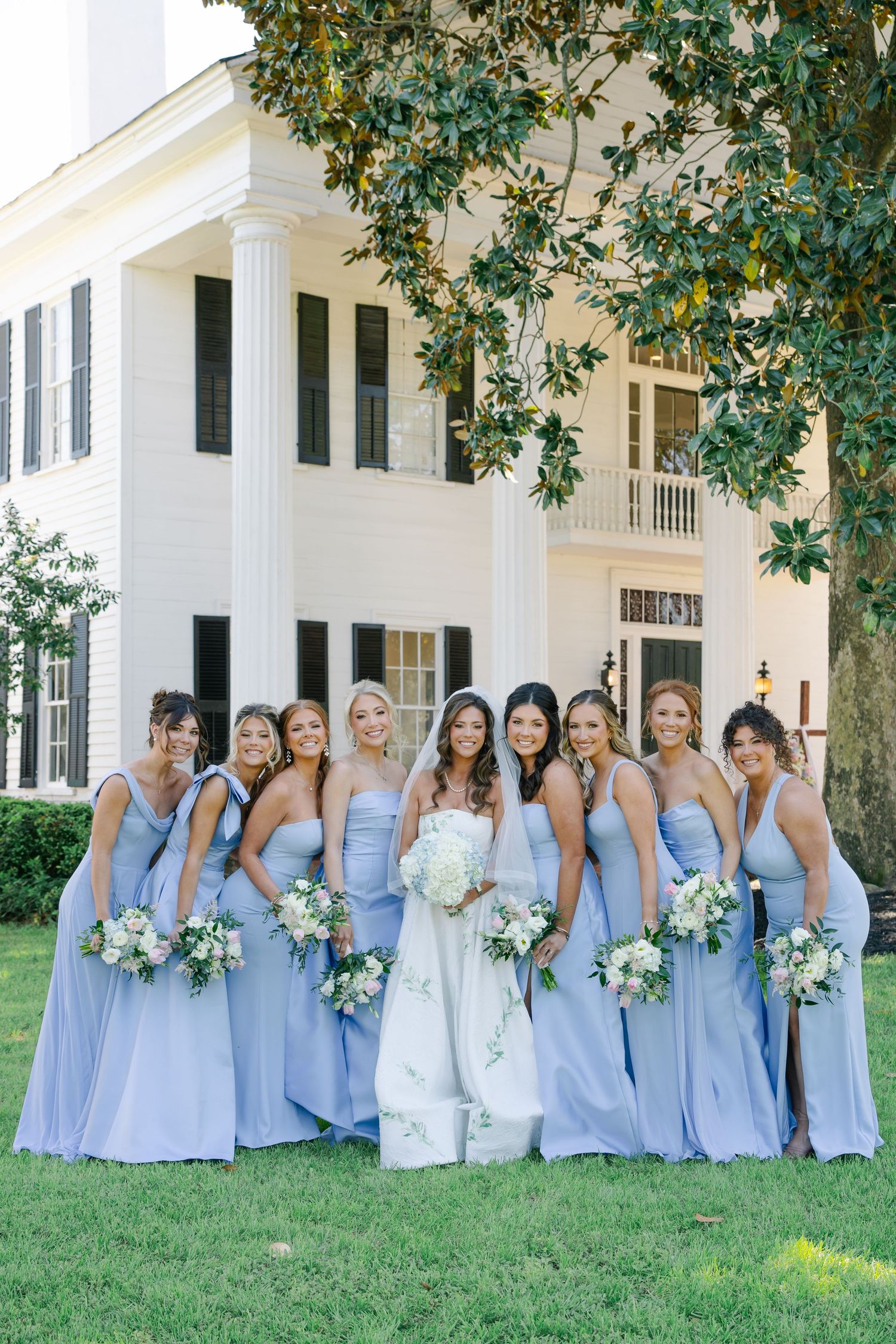 Smiling bride and bridesmaids in matching lavender blue gowns pose together on green lawn before a white colonial mansion.