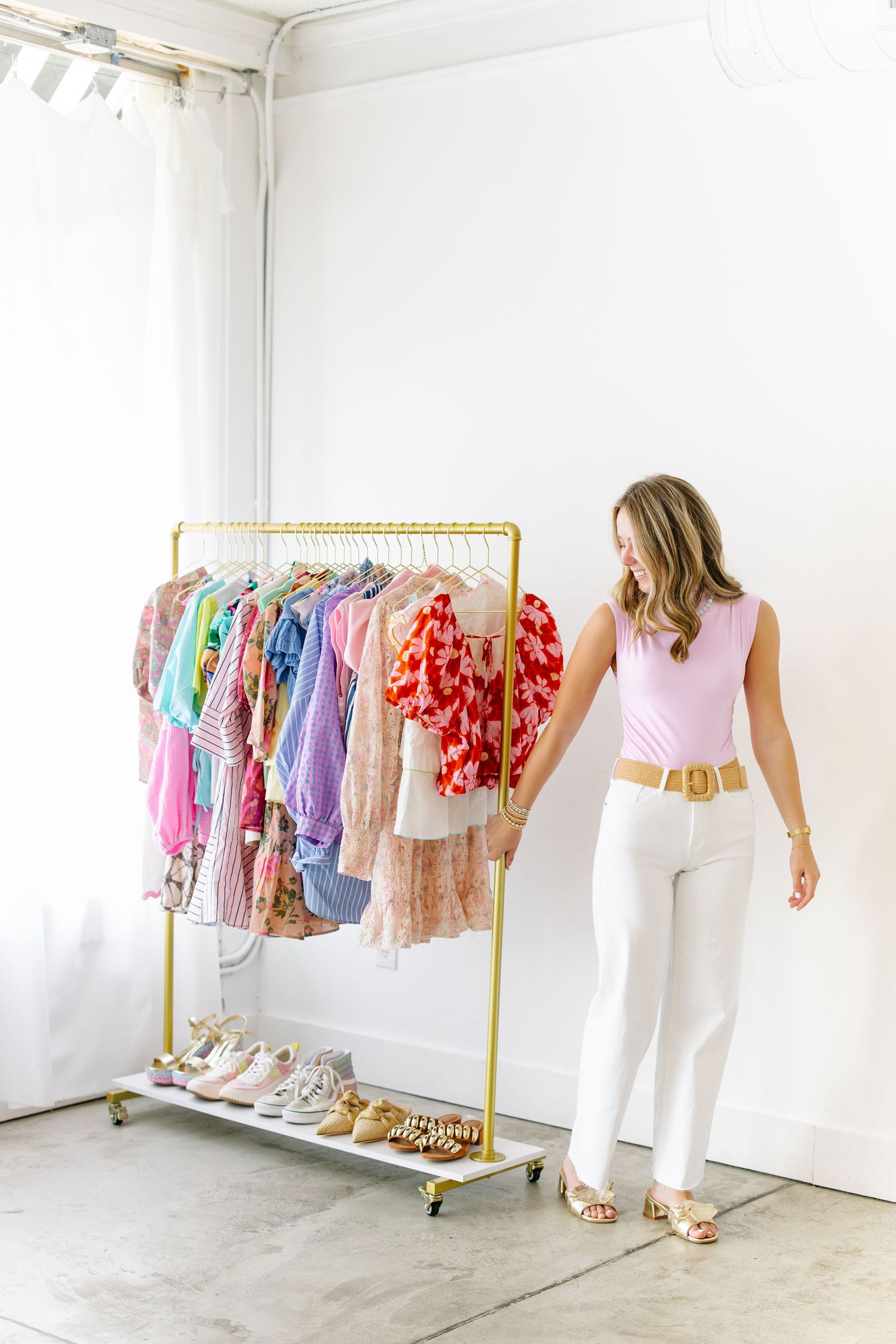 Blonde woman in pink top and white jeans standing next to gold rolling clothing rack with shoes below.