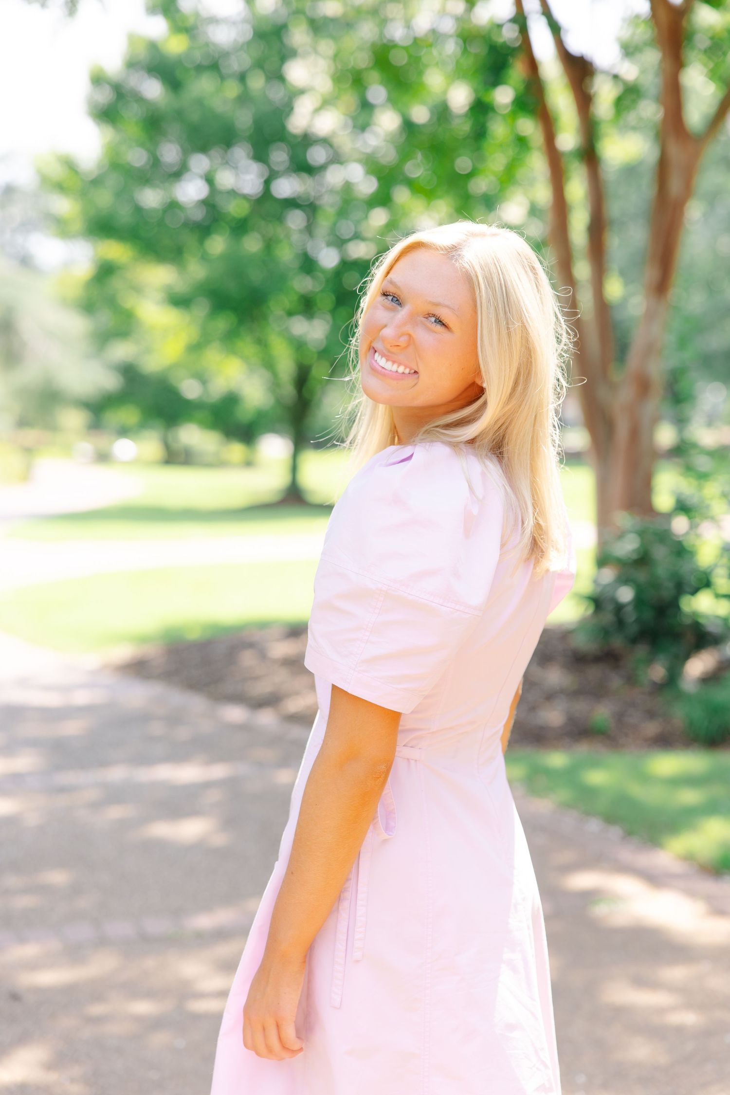 Laughing blonde woman in pink dress looking back over shoulder on sunny garden path.