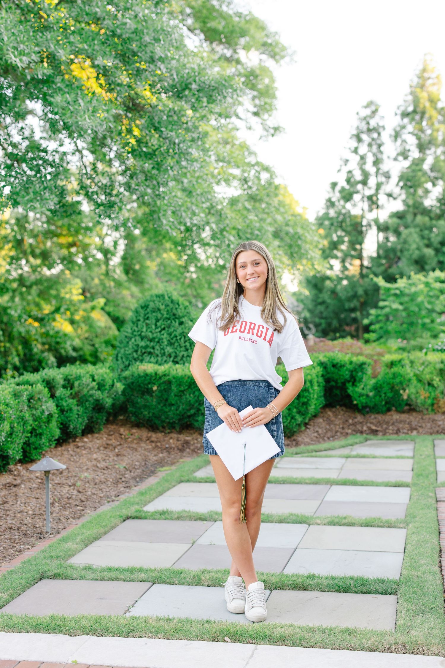 Smiling teen in Georgia Bulldogs shirt and denim skirt holding diploma on a garden stone pathway.