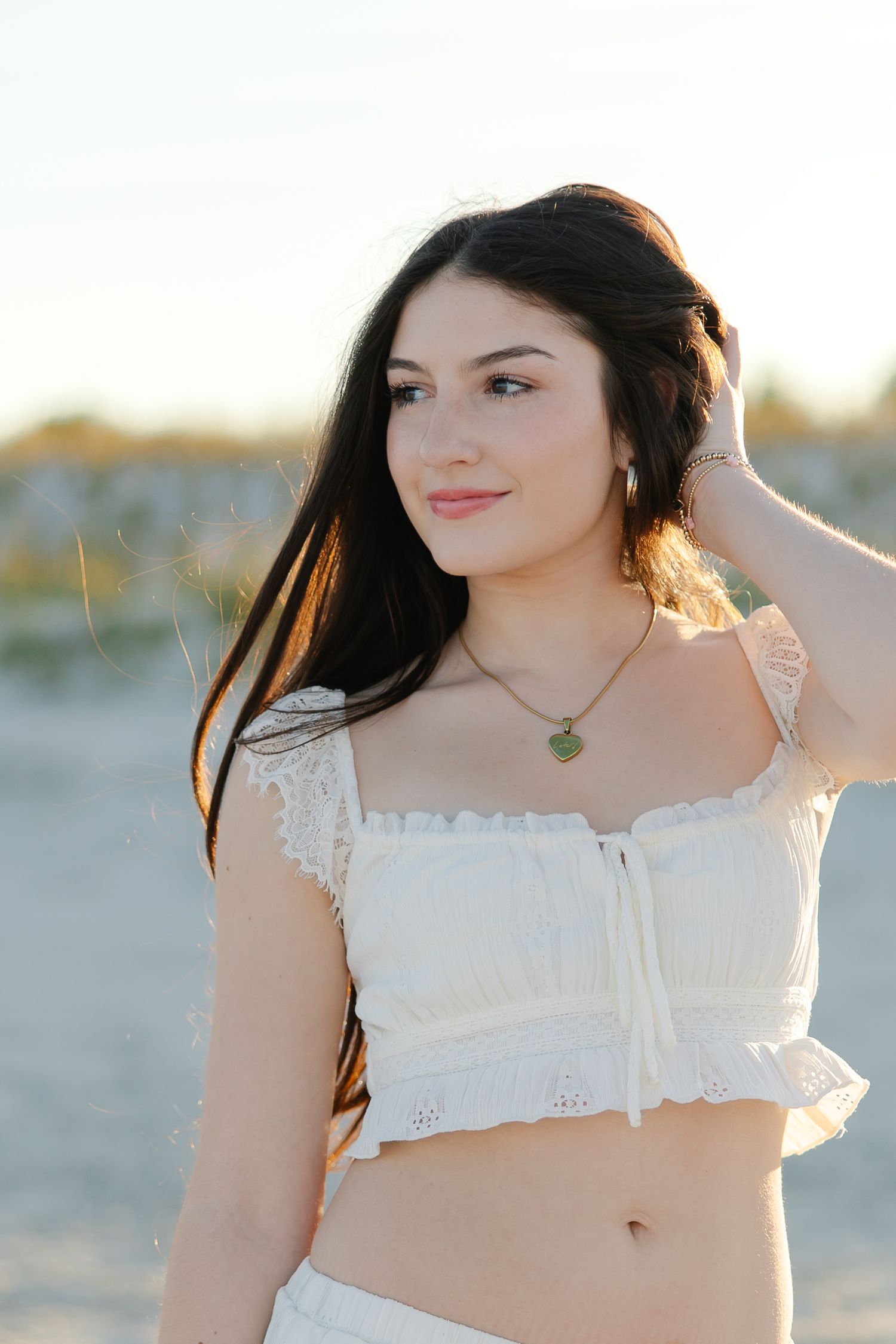 Young brunette woman in white lace crop top posing on sandy beach at golden hour.