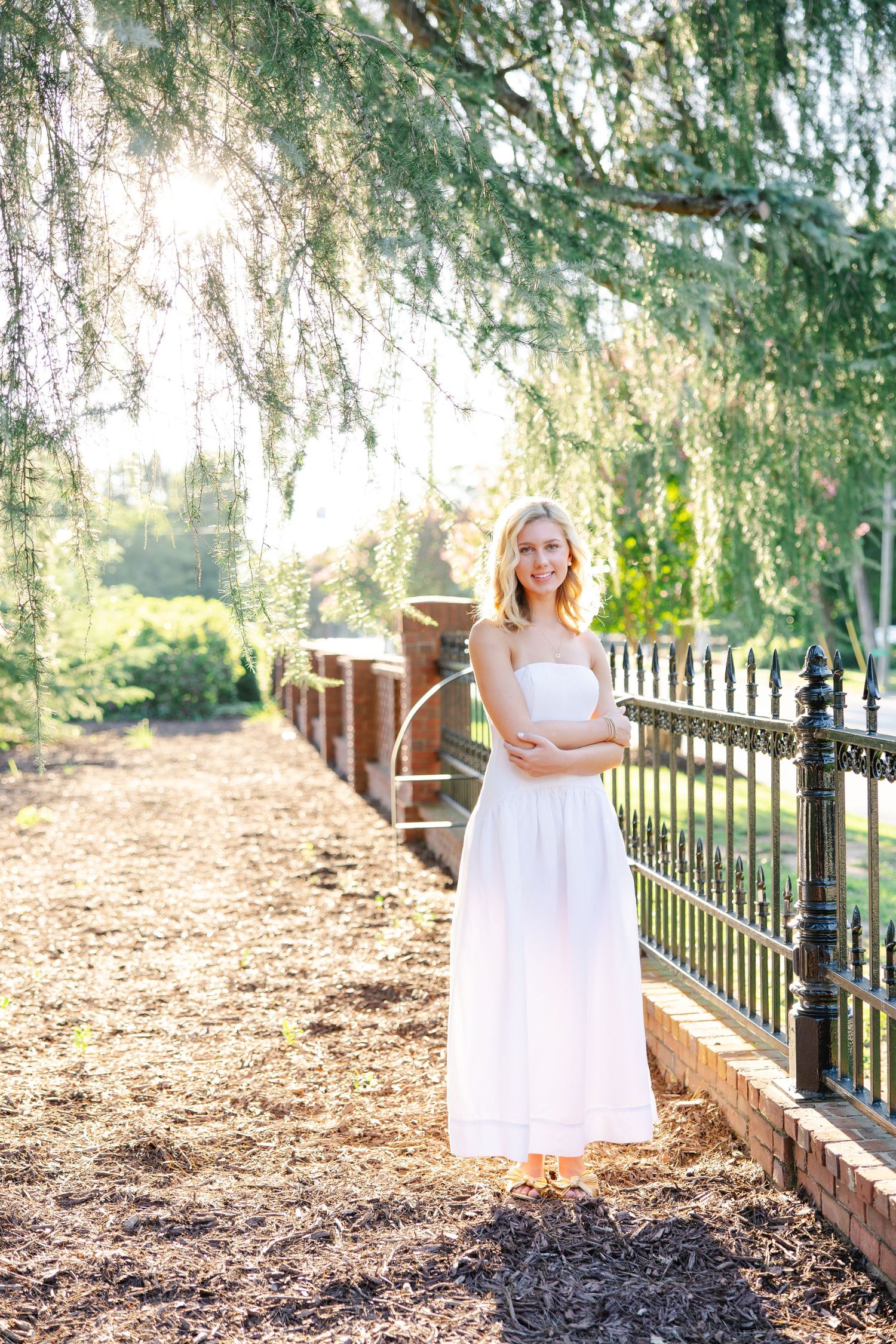 Young blonde woman in white maxi dress standing by iron fence in sunlit garden.