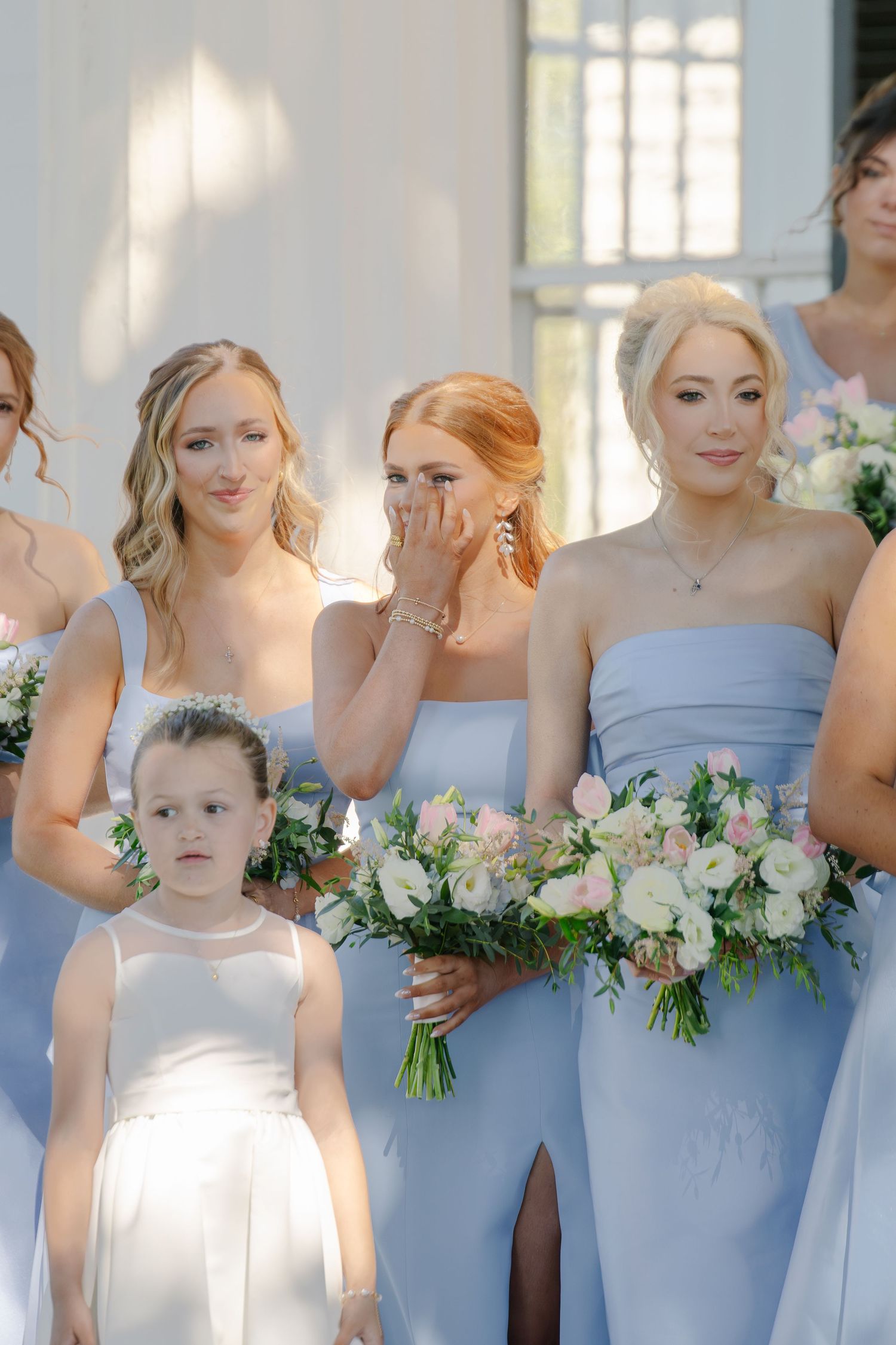 Bridesmaids in light blue gowns holding pink and white bouquets with emotional expressions.