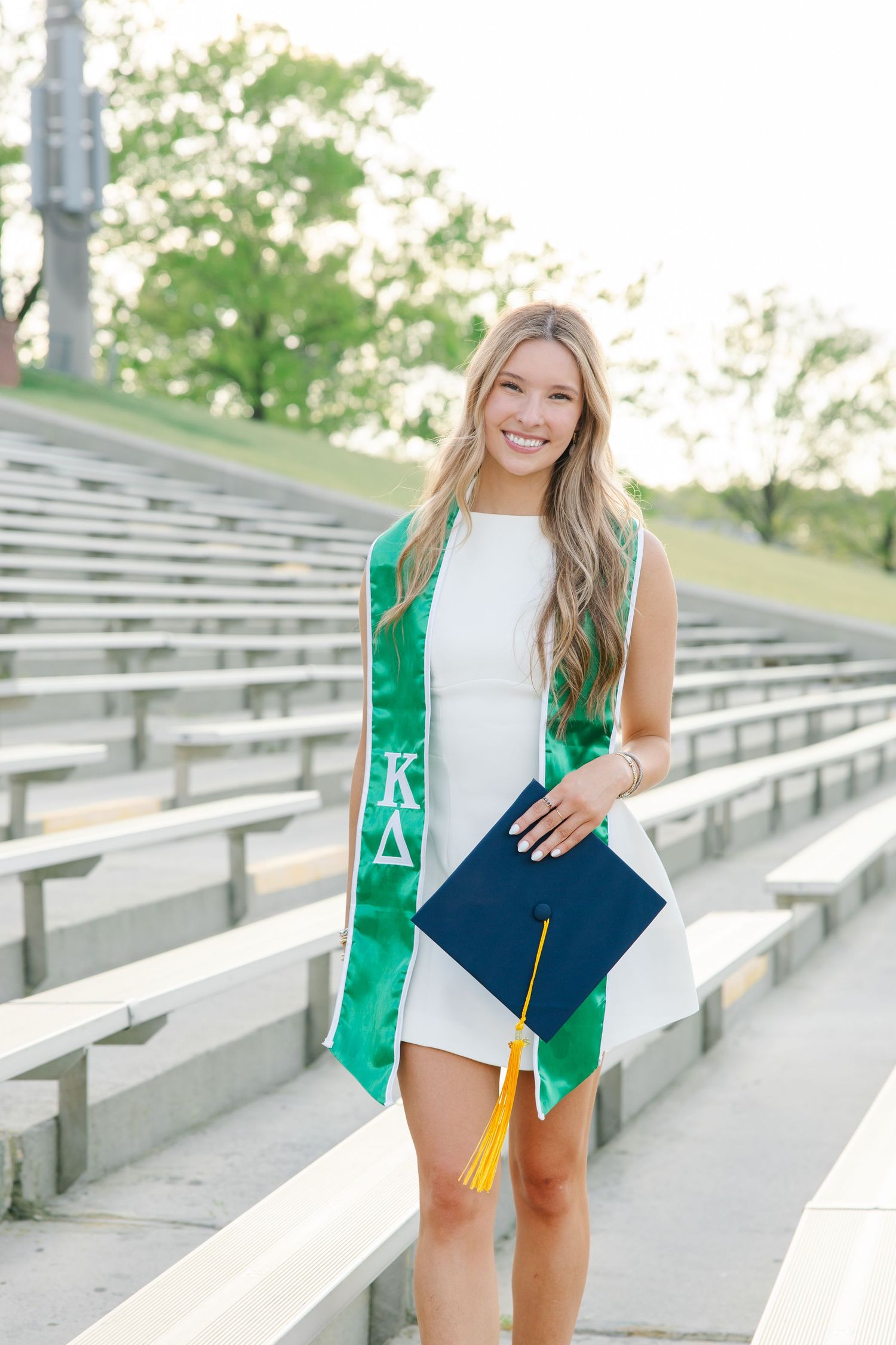 Female graduate in white dress holding navy cap wearing Kappa Delta green sorority graduation stole.