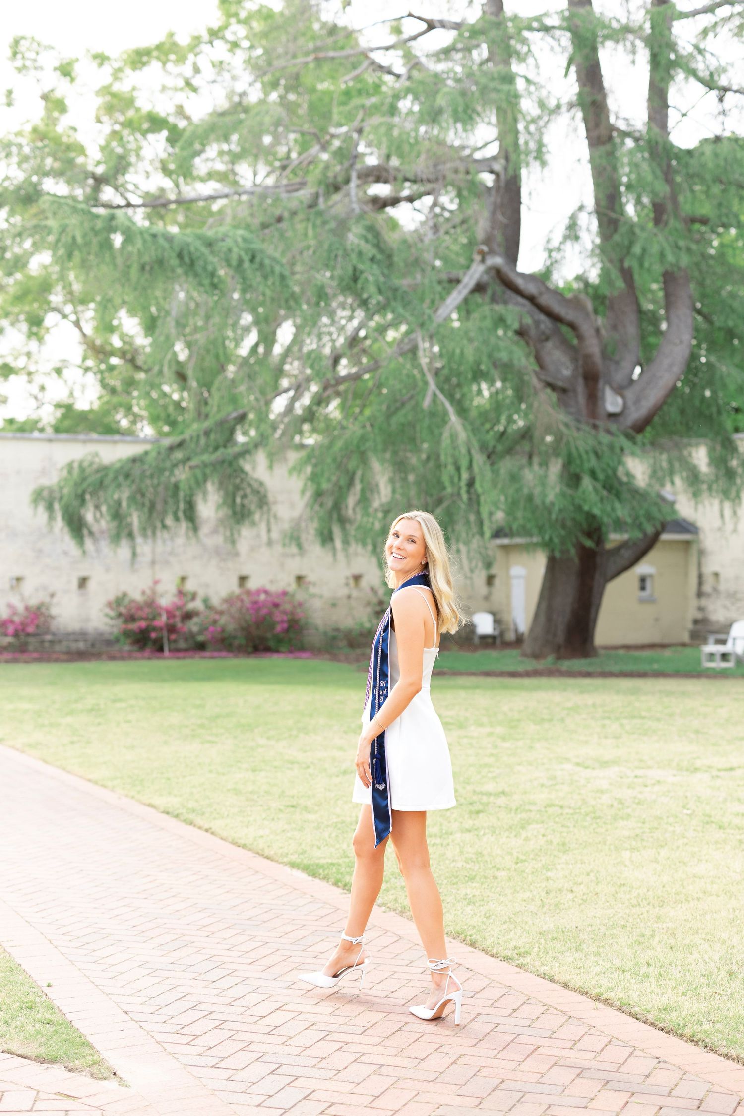 Blonde graduate in white mini dress with navy stole poses on brick path with green lawn.