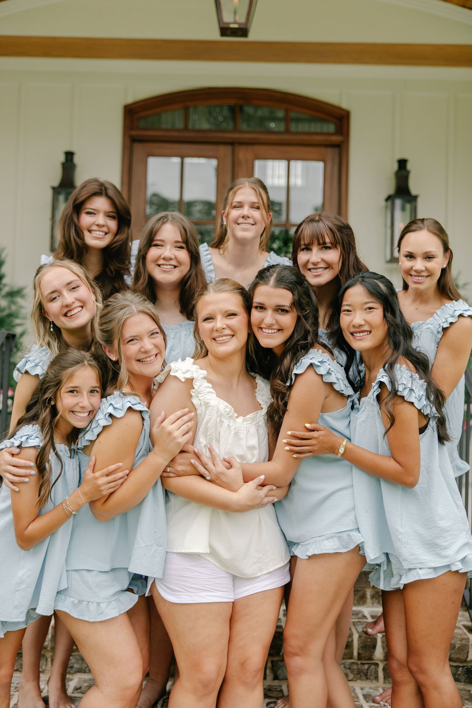 Bride surrounded by smiling bridesmaids in light blue matching sets posing on home steps before wedding.