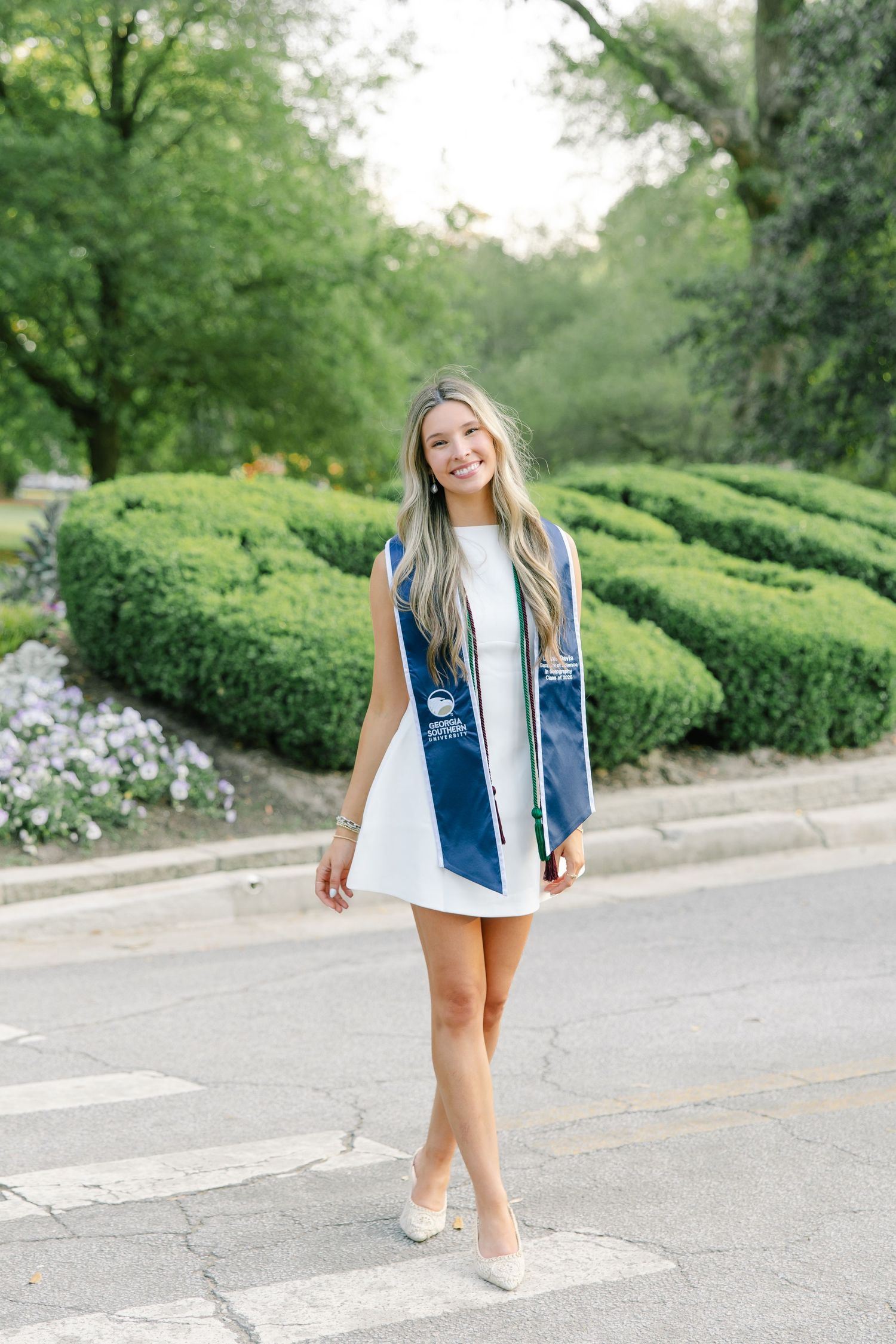 Female graduate in white dress with Georgia Southern blue stole walking near campus garden path.