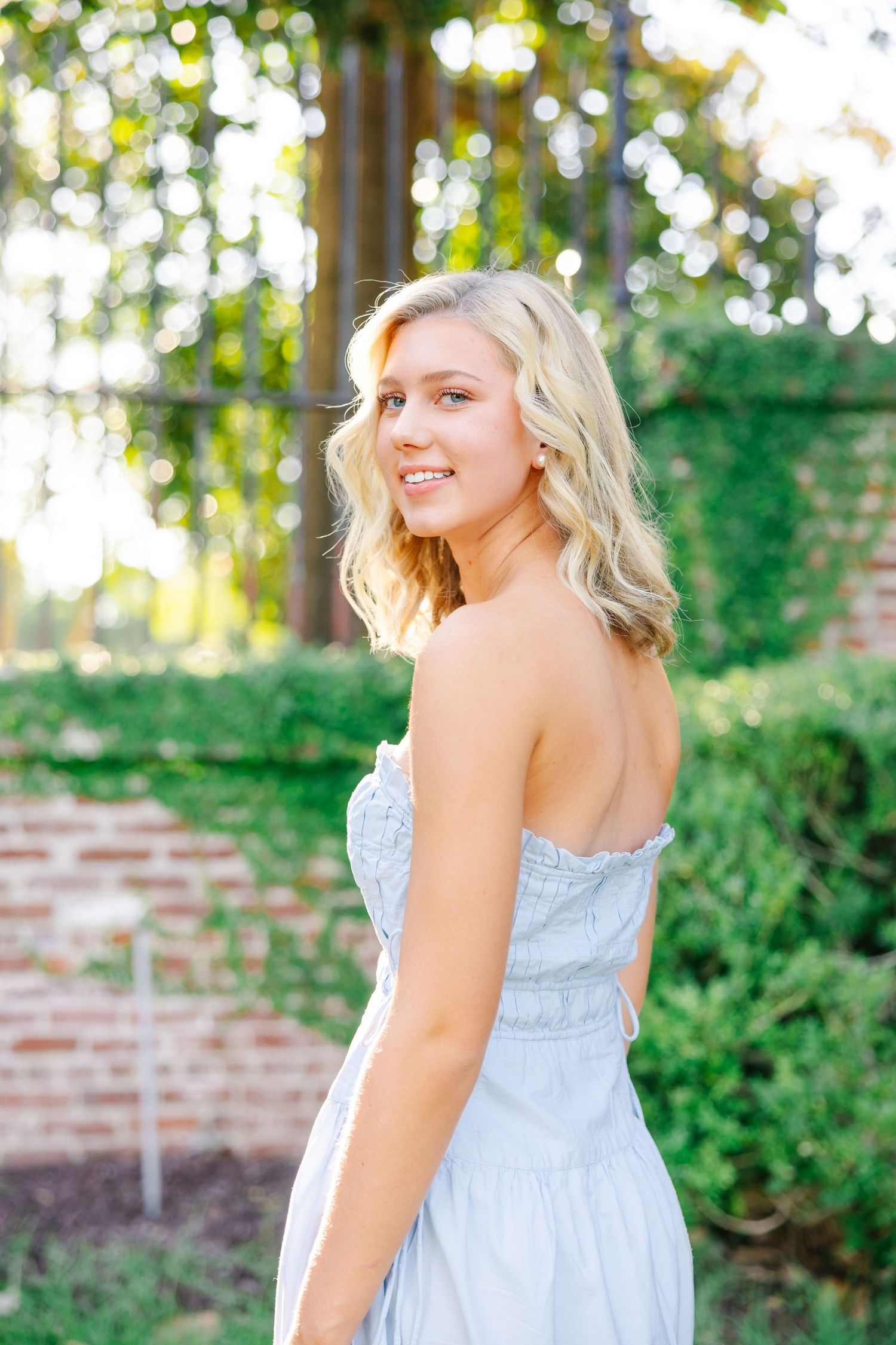 Smiling blonde woman in strapless light blue dress standing in lush garden setting.