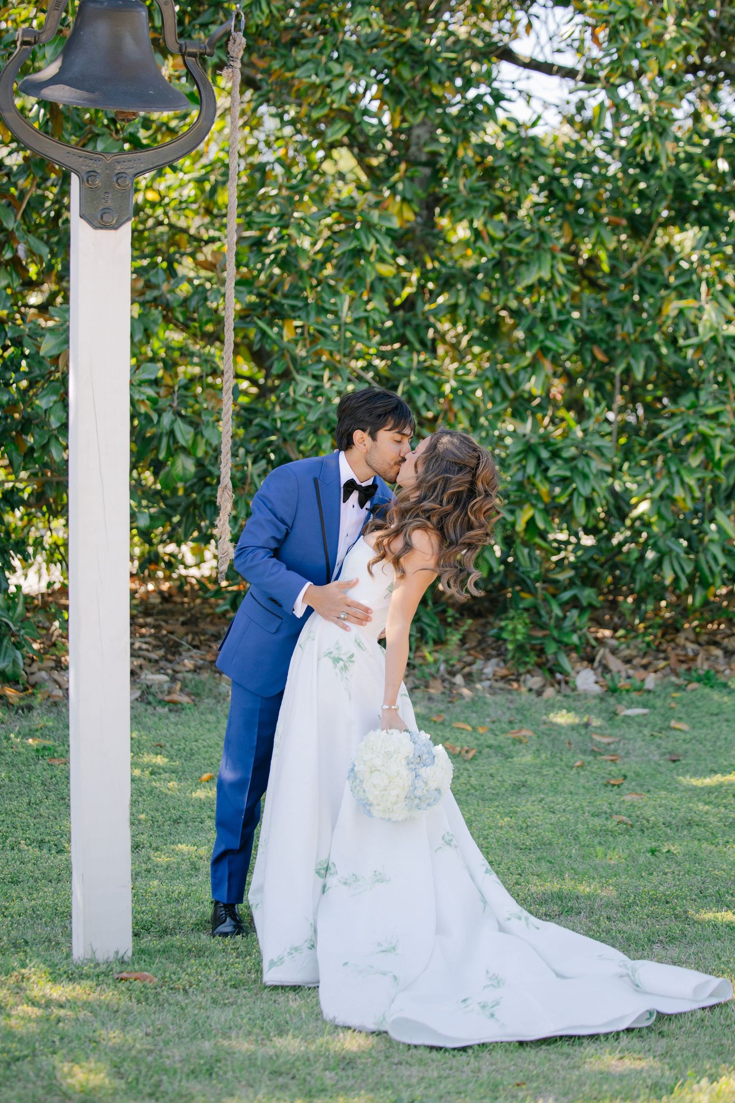 Bride and groom sharing a romantic kiss outdoors near a bronze bell, bride holding white hydrangea bouquet in garden setting.