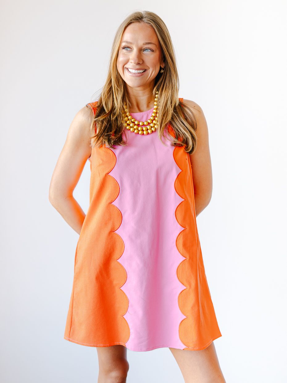 Woman wearing a pink and orange colorblock scalloped sleeveless shift dress with yellow bead necklace.