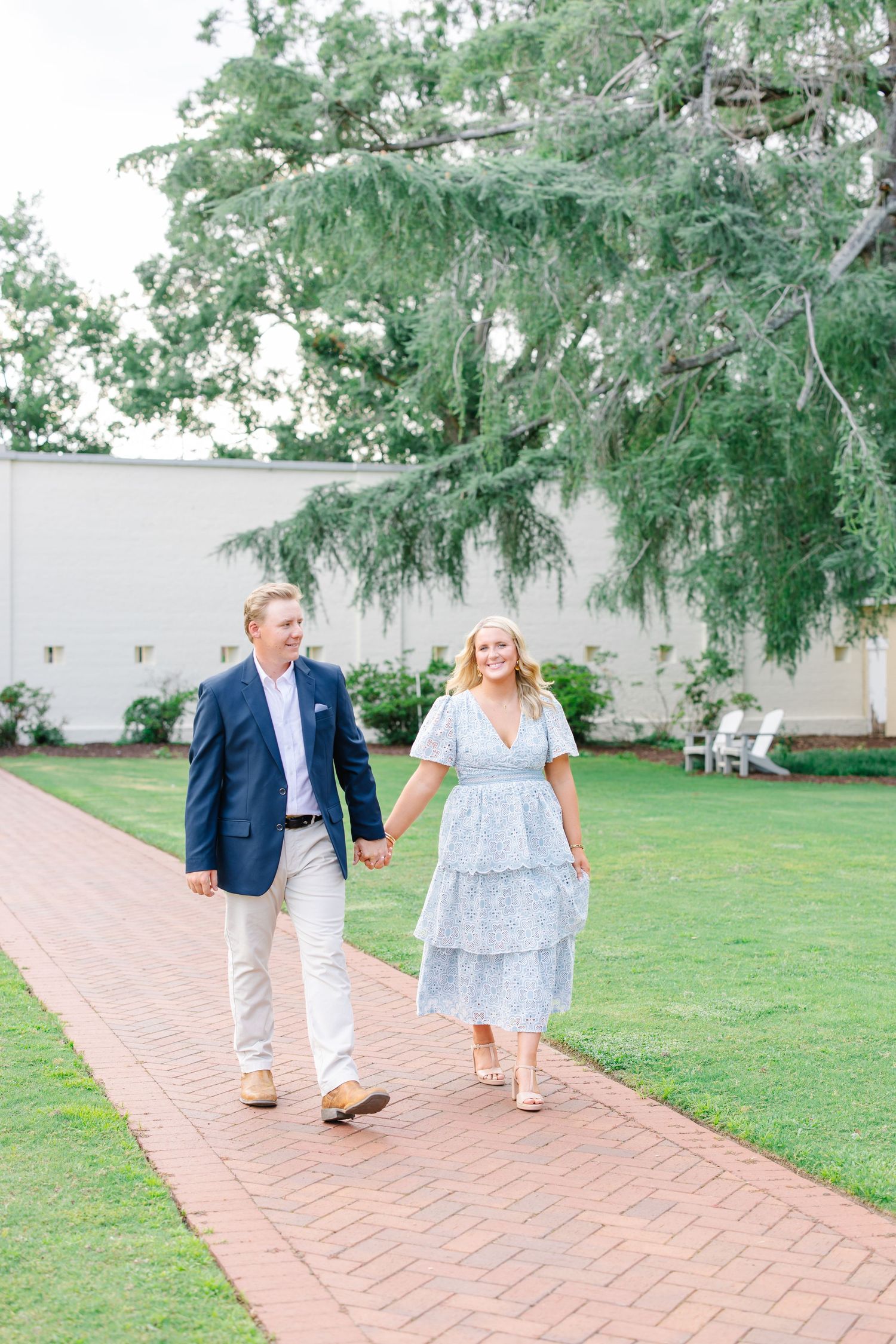 Happy couple strolling hand in hand along brick path beneath large weeping tree.