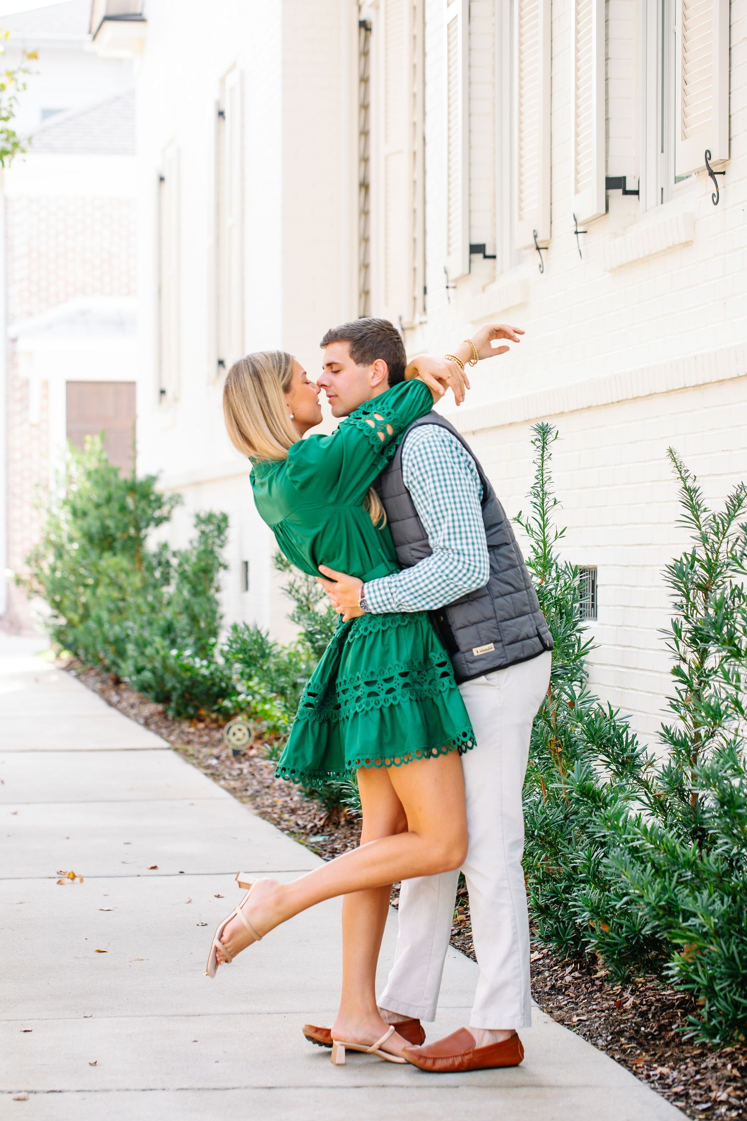 Couple sharing a kiss on sidewalk beside white building with green shrubs nearby.