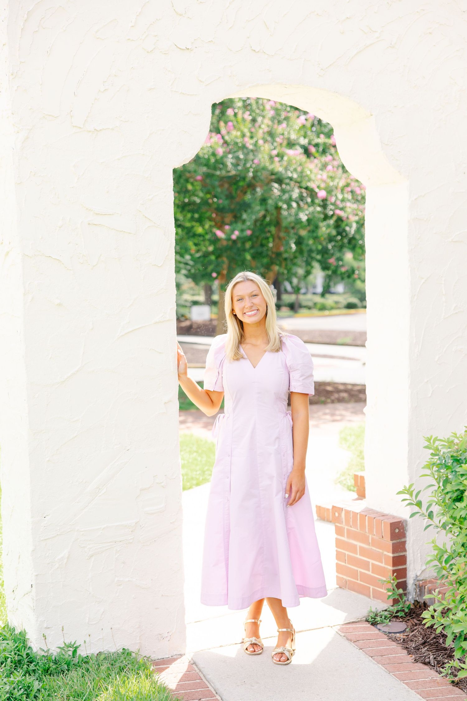 Smiling blonde woman in pink dress posing inside white stucco archway outdoors.