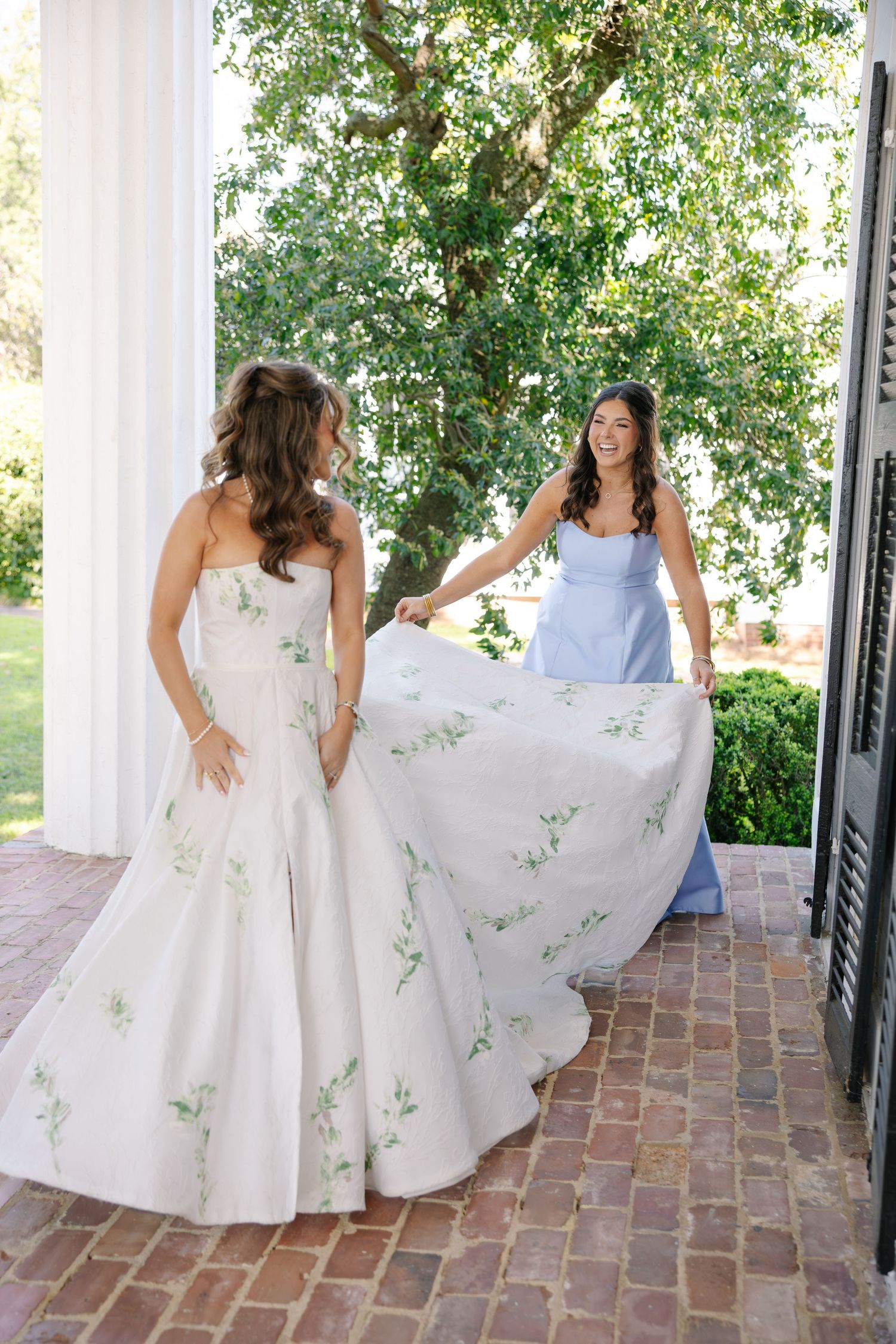 Bridesmaid in periwinkle blue dress helps lift the floral embroidered train of the bride's elegant white wedding gown.