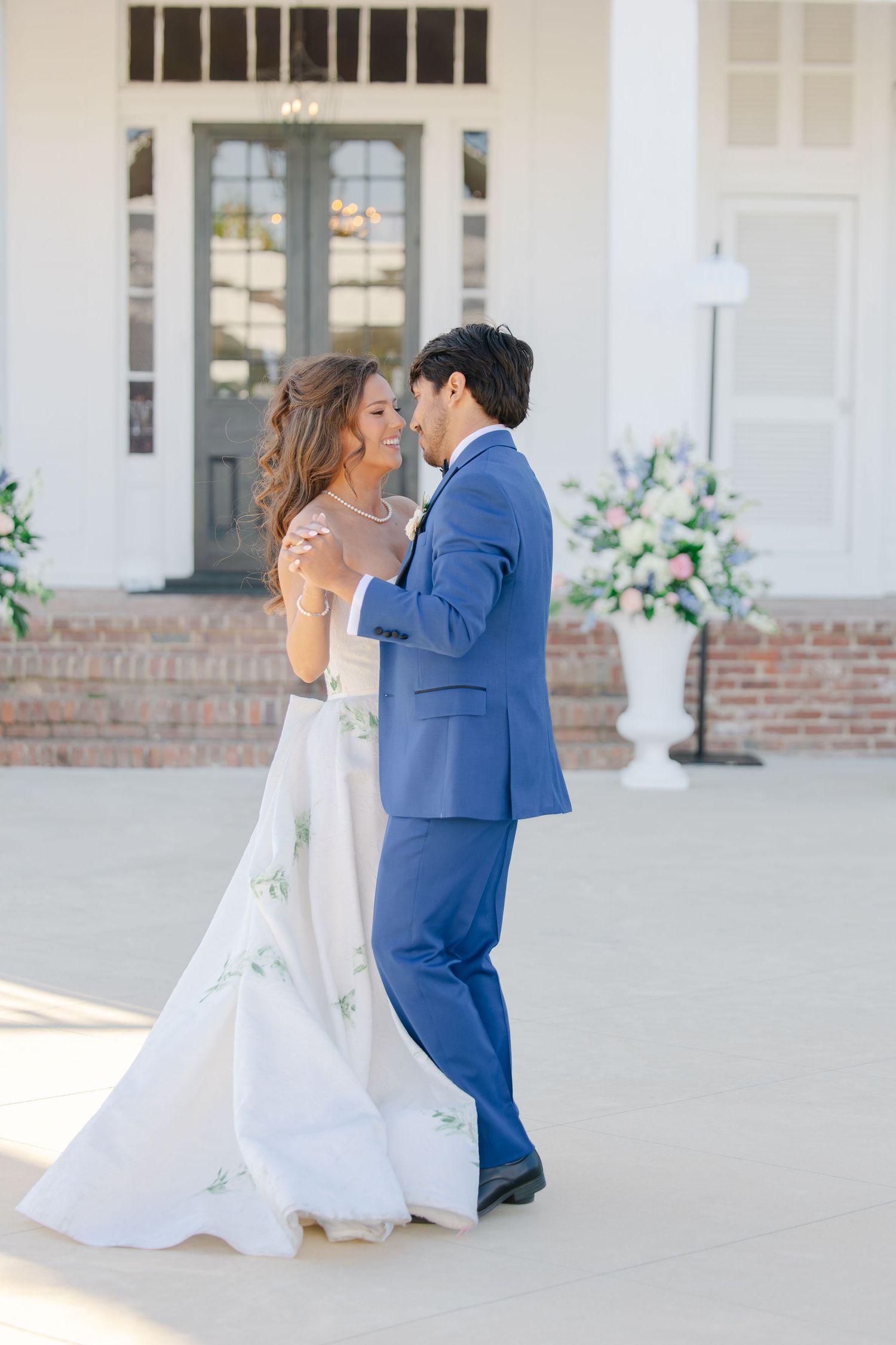 Bride in floral embroidered gown dancing with groom in blue tuxedo outside elegant white venue.