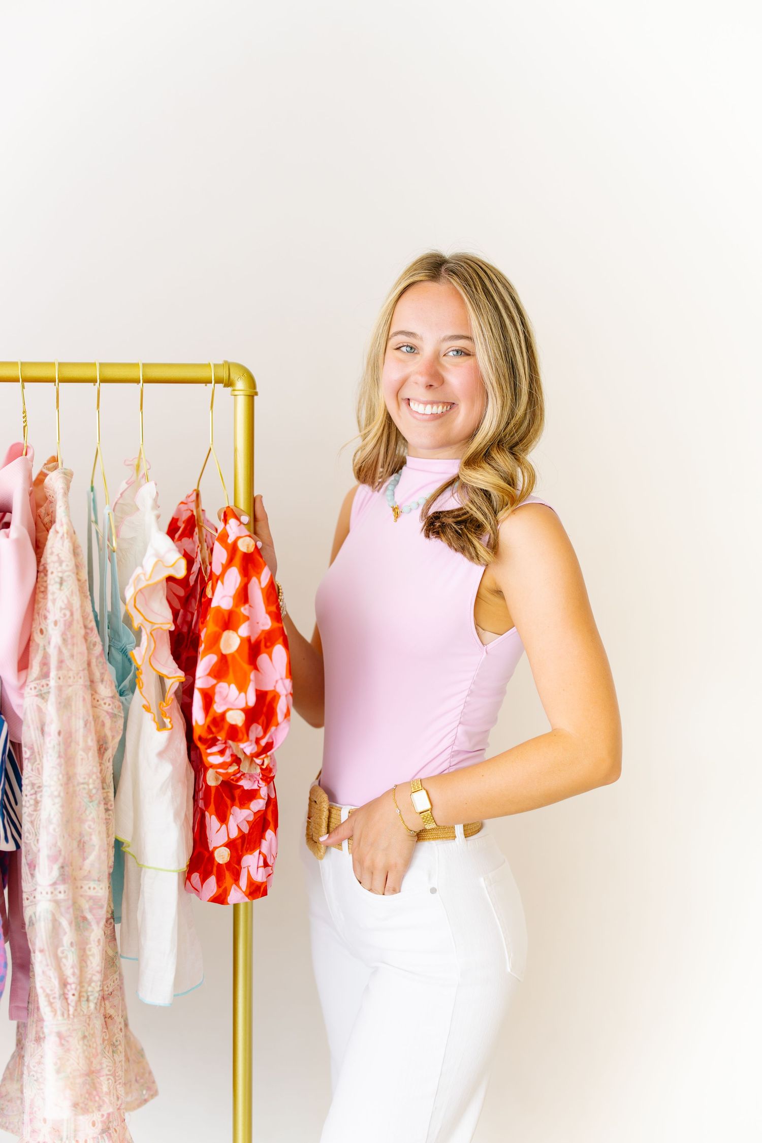 Cheerful blonde woman in pink top and white pants holding a red floral dress beside a clothing rack.