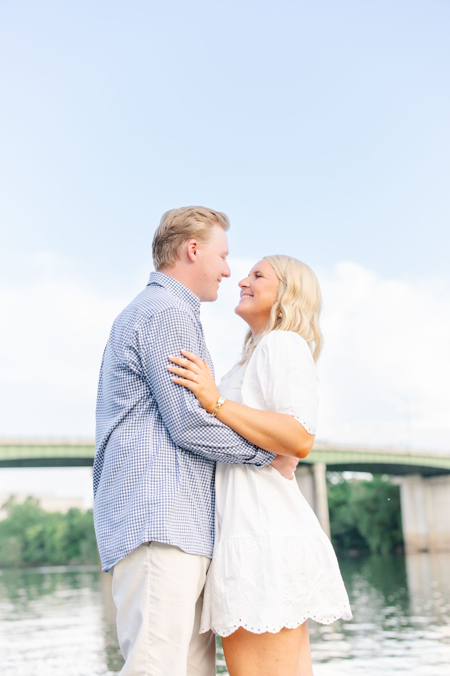 Couple embracing and smiling at each other with river and bridge in background.
