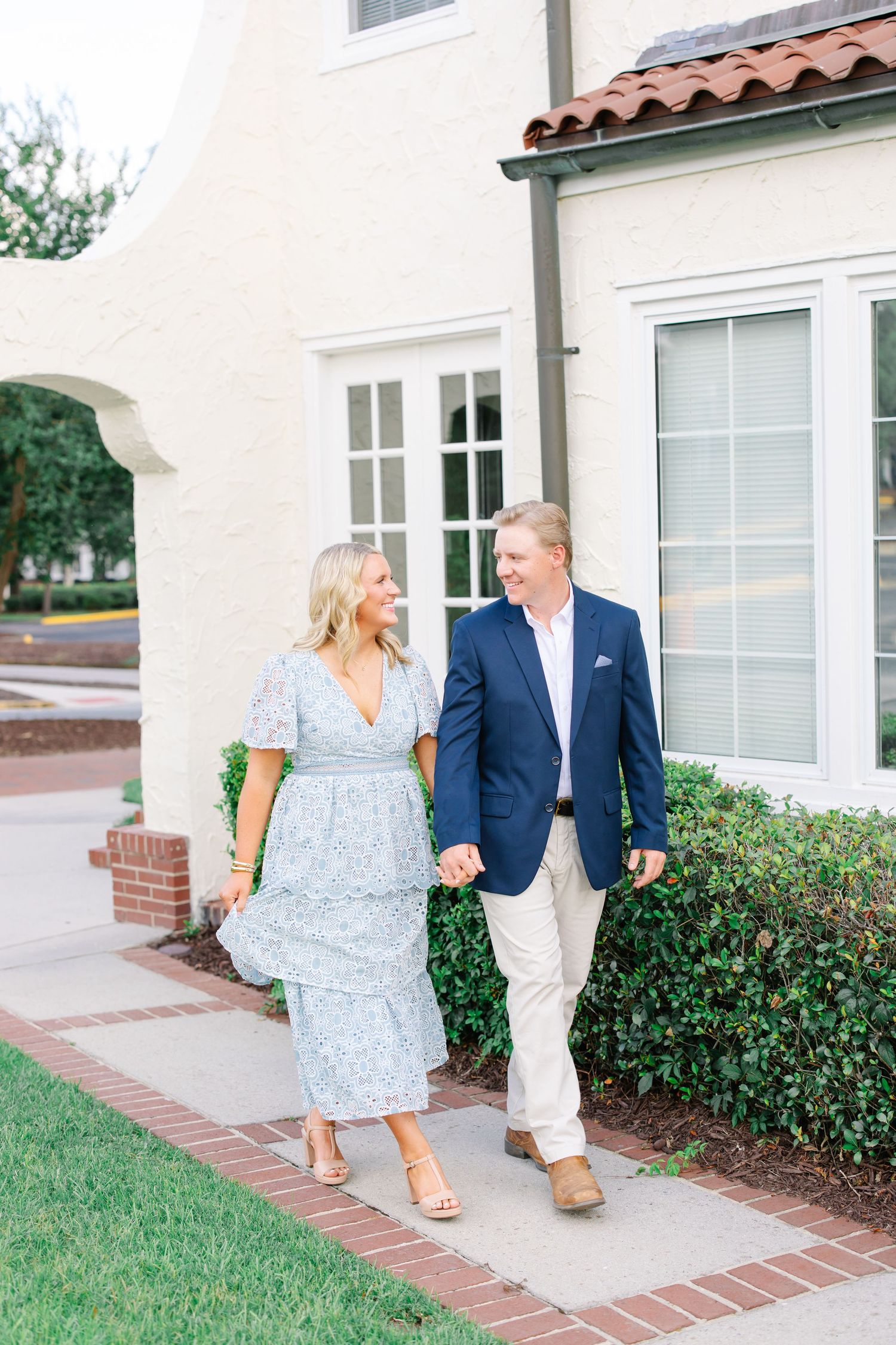 Smiling couple walking hand in hand past white stucco building with lush green hedges.