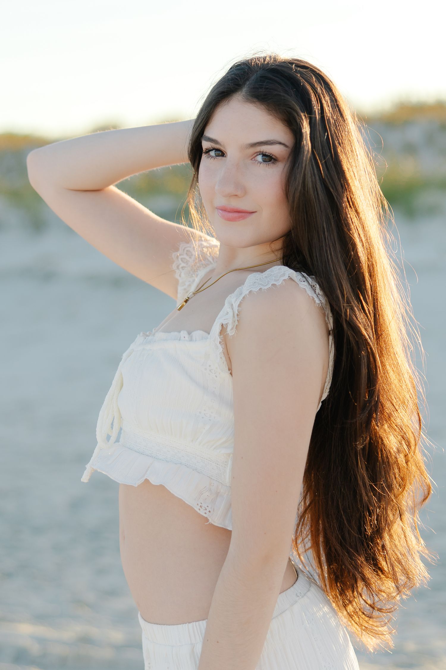 Woman with long dark hair in white crop top standing on sandy beach at sunset.