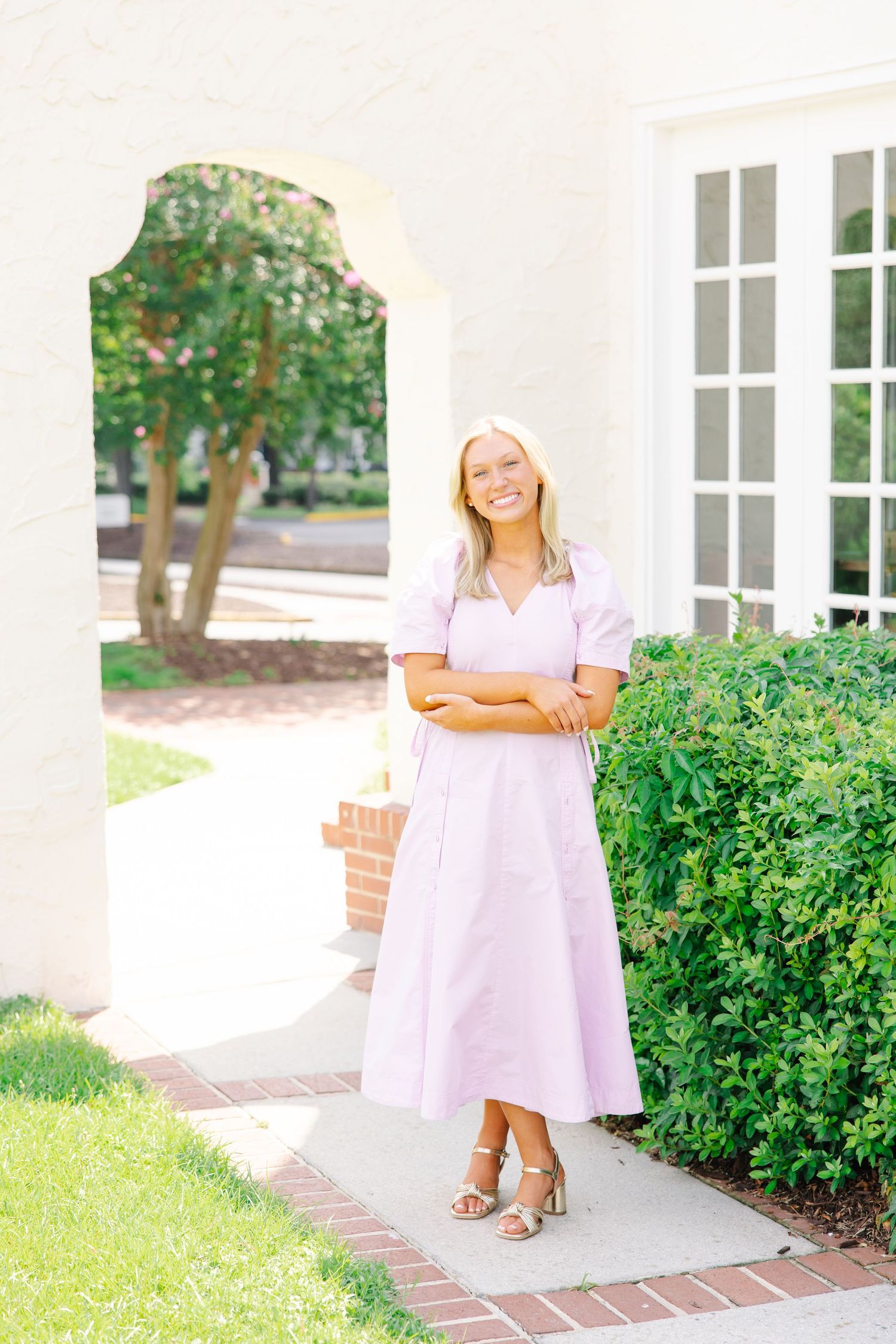 Blonde woman in lavender midi dress standing under white arch beside green bushes.