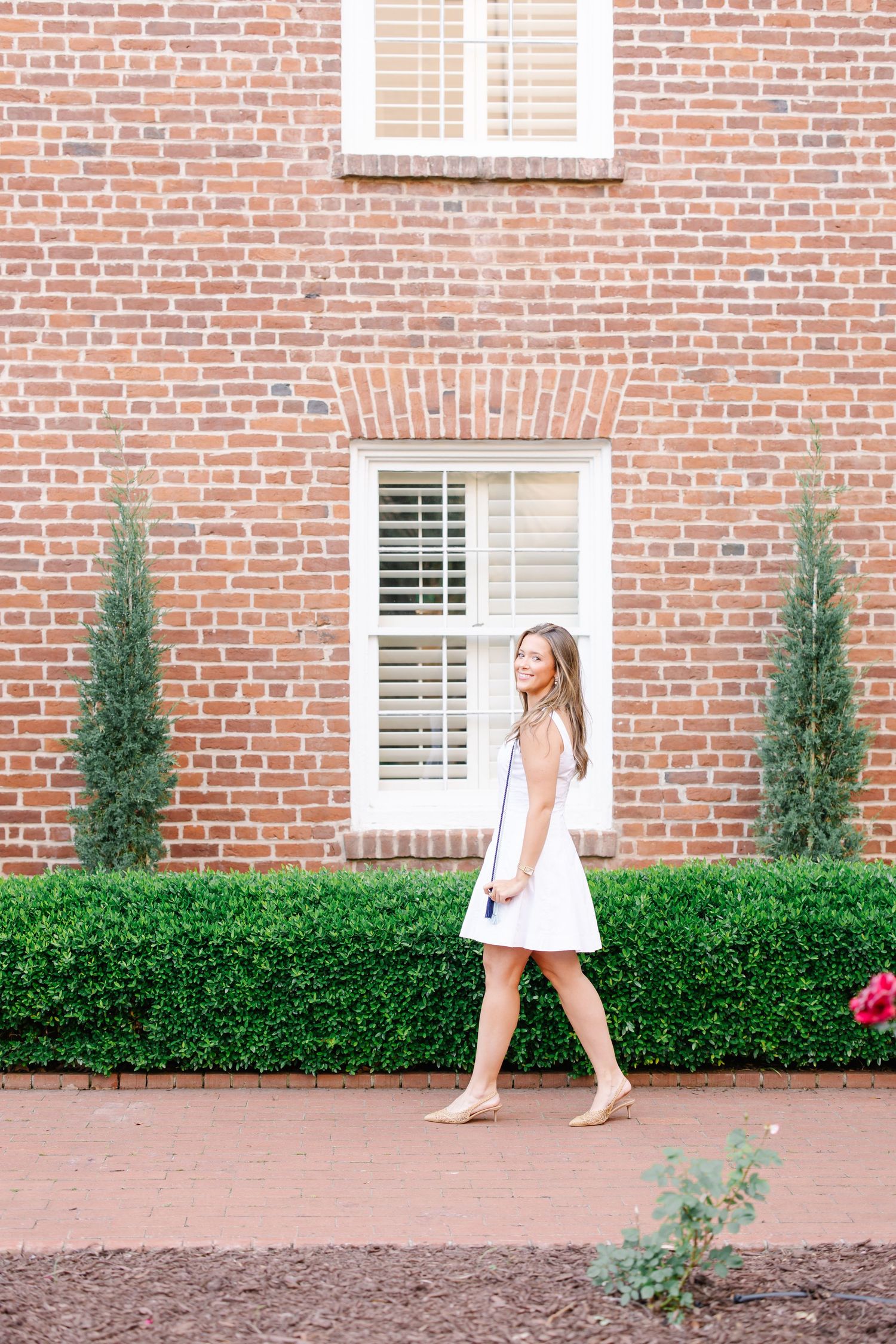 Young woman in white dress walking along brick building with manicured hedges.